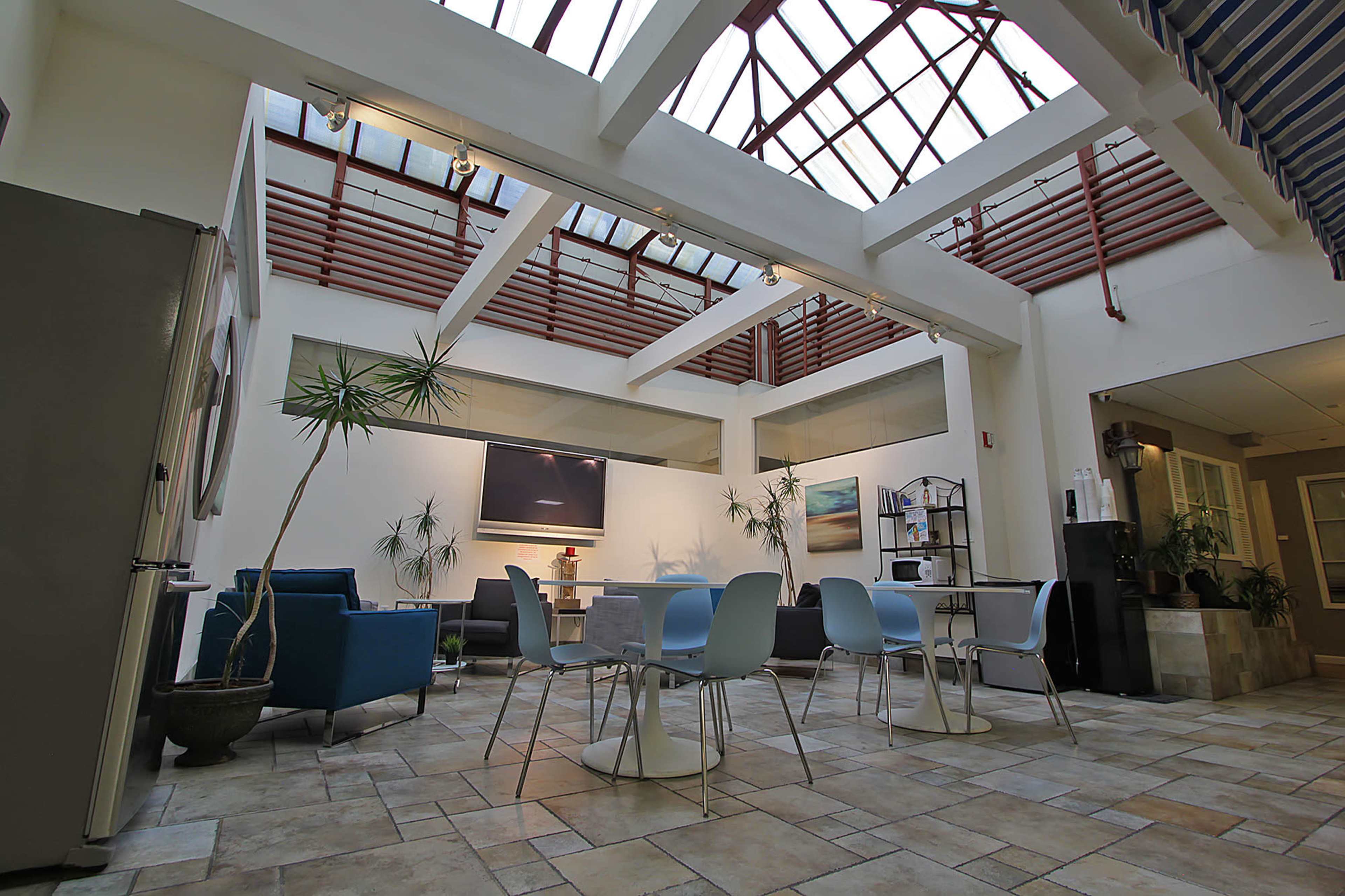 The image shows a modern, open interior space with a glass skylight, featuring seating areas, tables, and indoor plants.