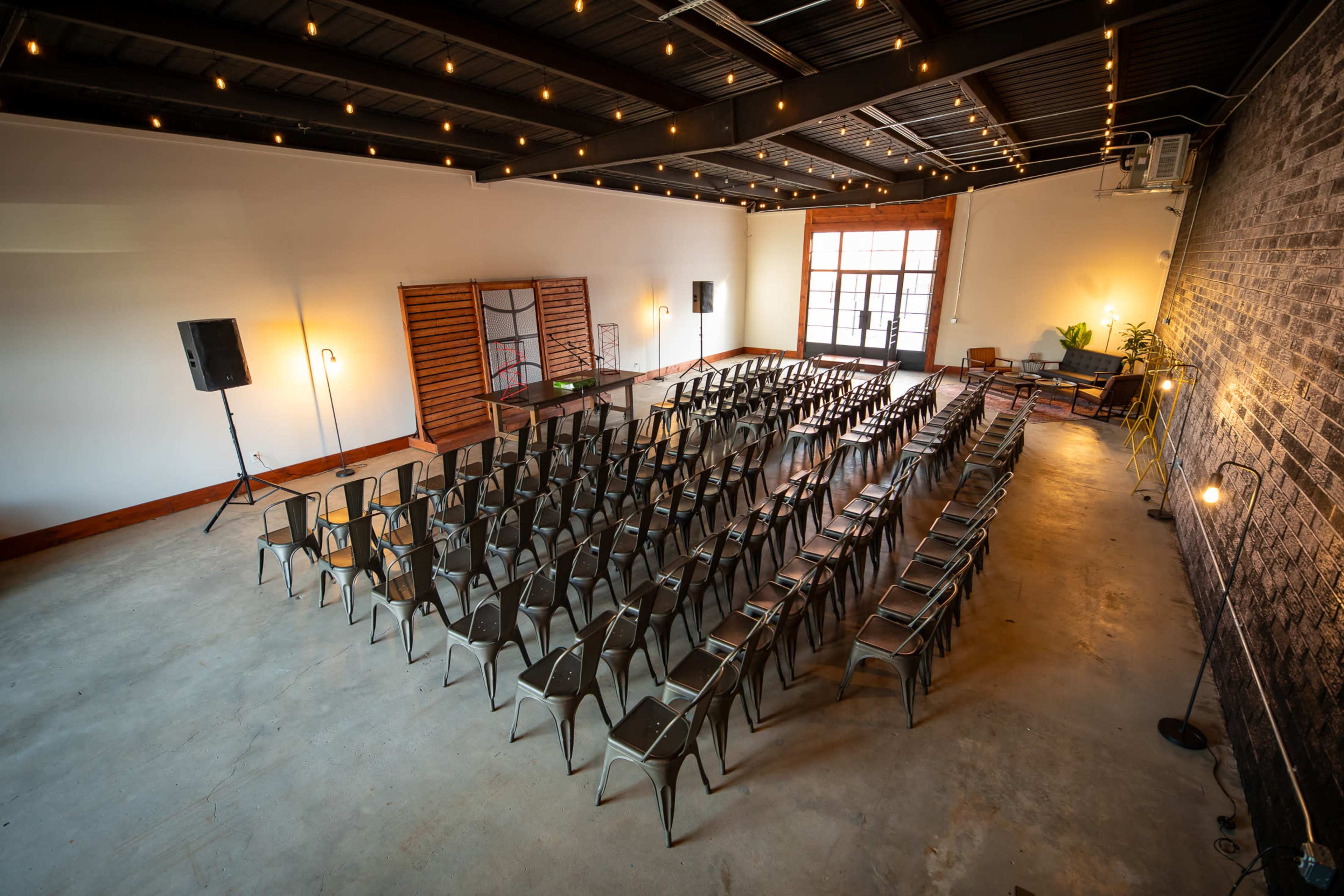 A spacious event venue features rows of metal chairs arranged for a gathering, with string lights illuminating the ceiling.