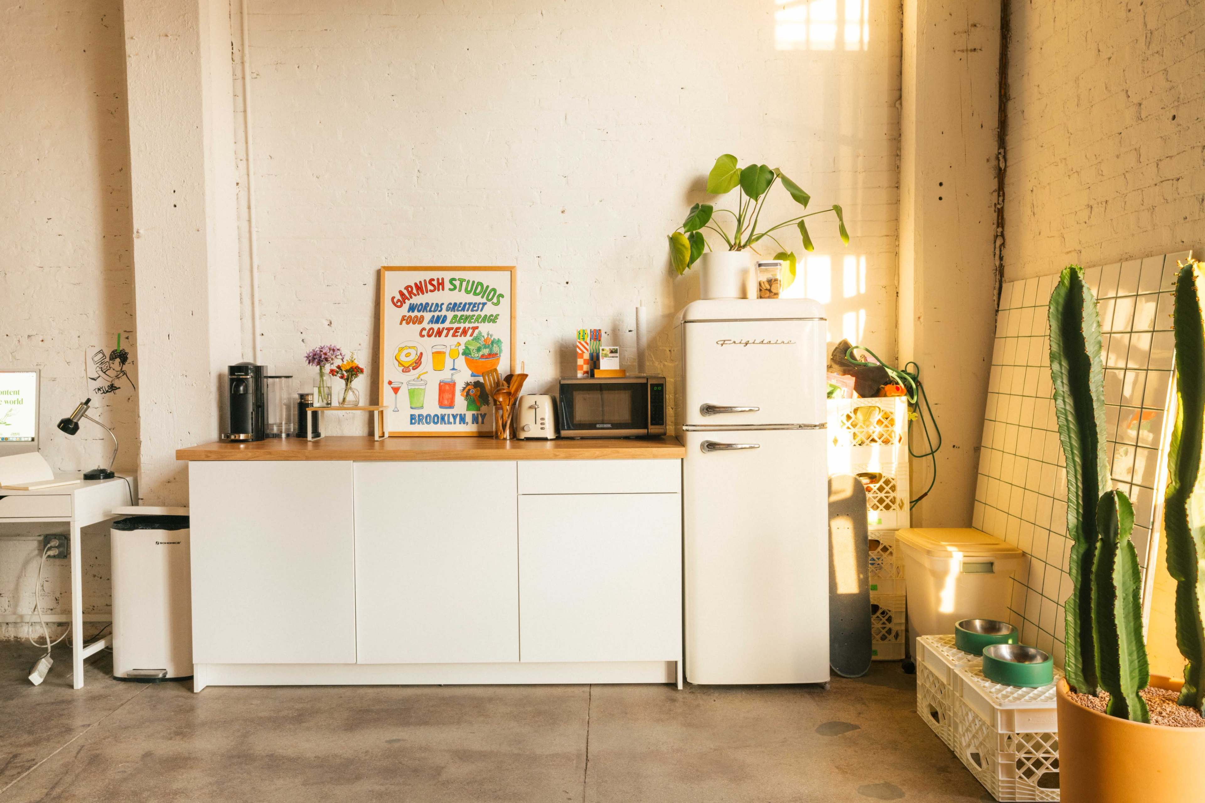 A bright kitchen featuring a white cabinet, a vintage-style refrigerator, and a colorful poster on the wall, with a cactus and a potted plant nearby.