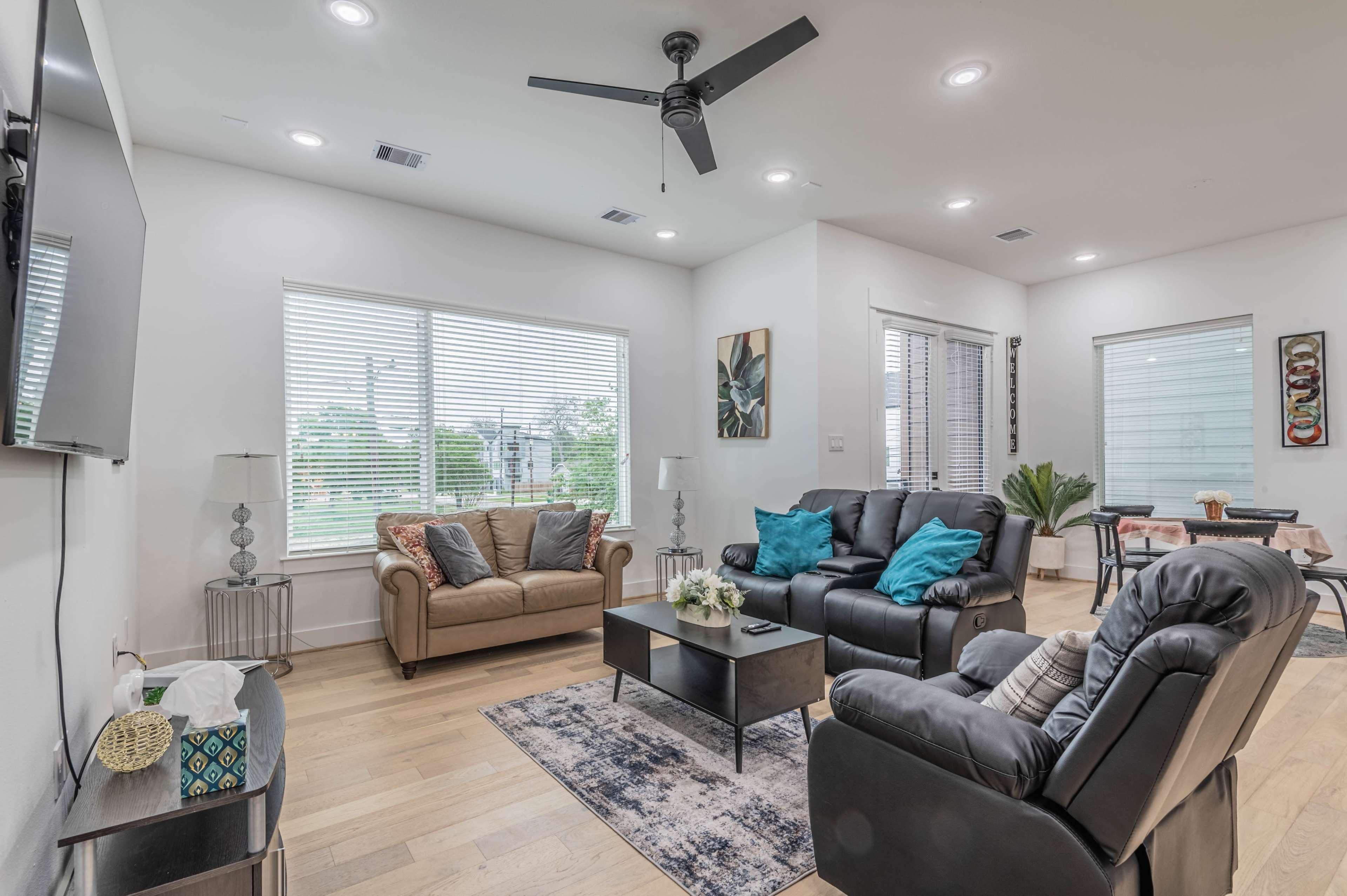 A modern living room with two sofas, a coffee table, and a TV mounted on the wall, overlooking windows with blinds.
