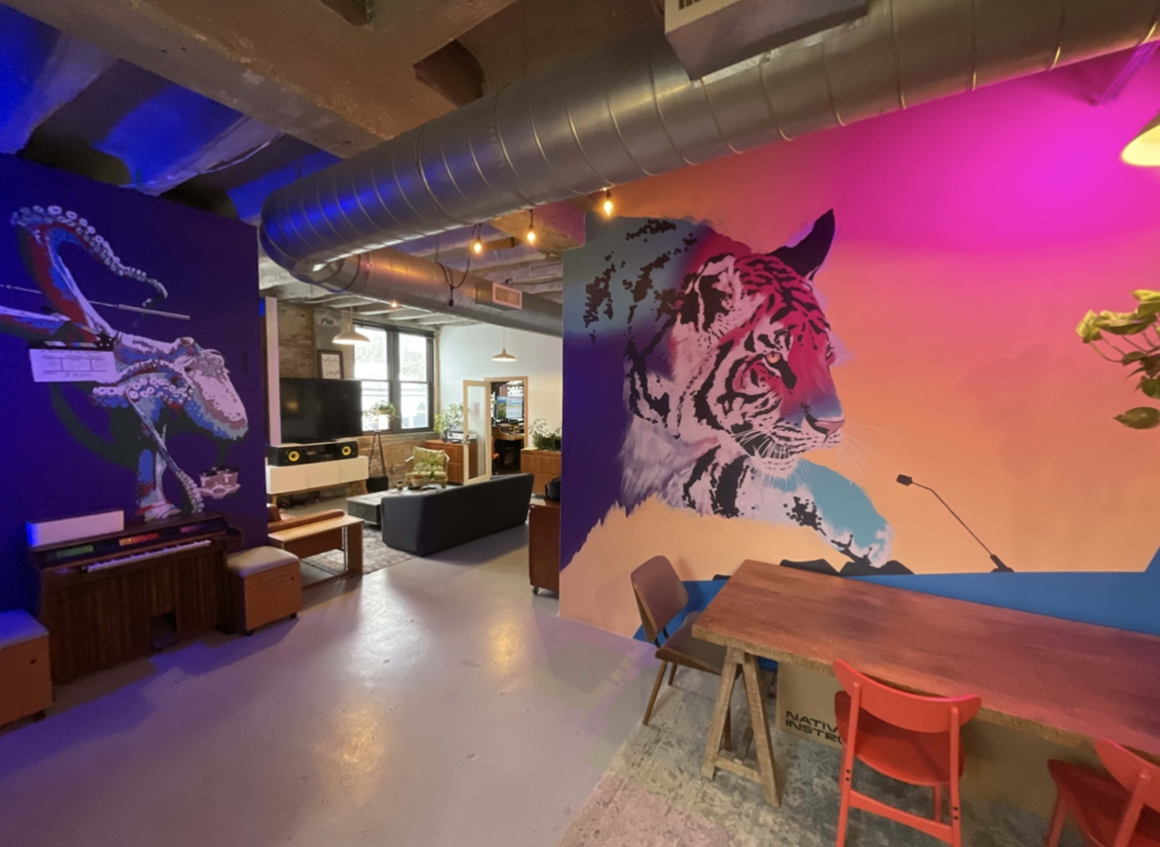 The image shows a modern, artistic communal space featuring vibrant wall murals of a tiger and an octopus, with furniture including a dining table, chairs, and a couch.
