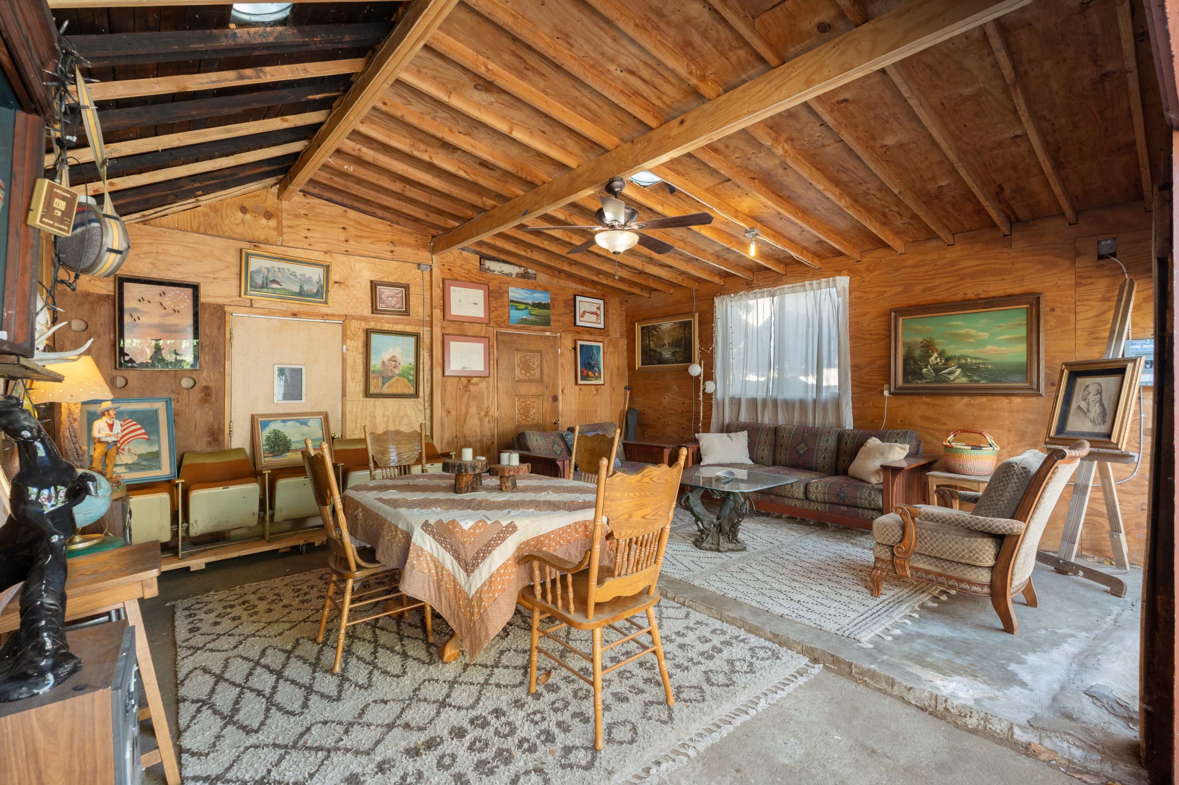 The interior of a rustic cabin features a dining area with a round table and several wooden chairs, adorned with various artworks on the walls and a patterned rug covering the floor.