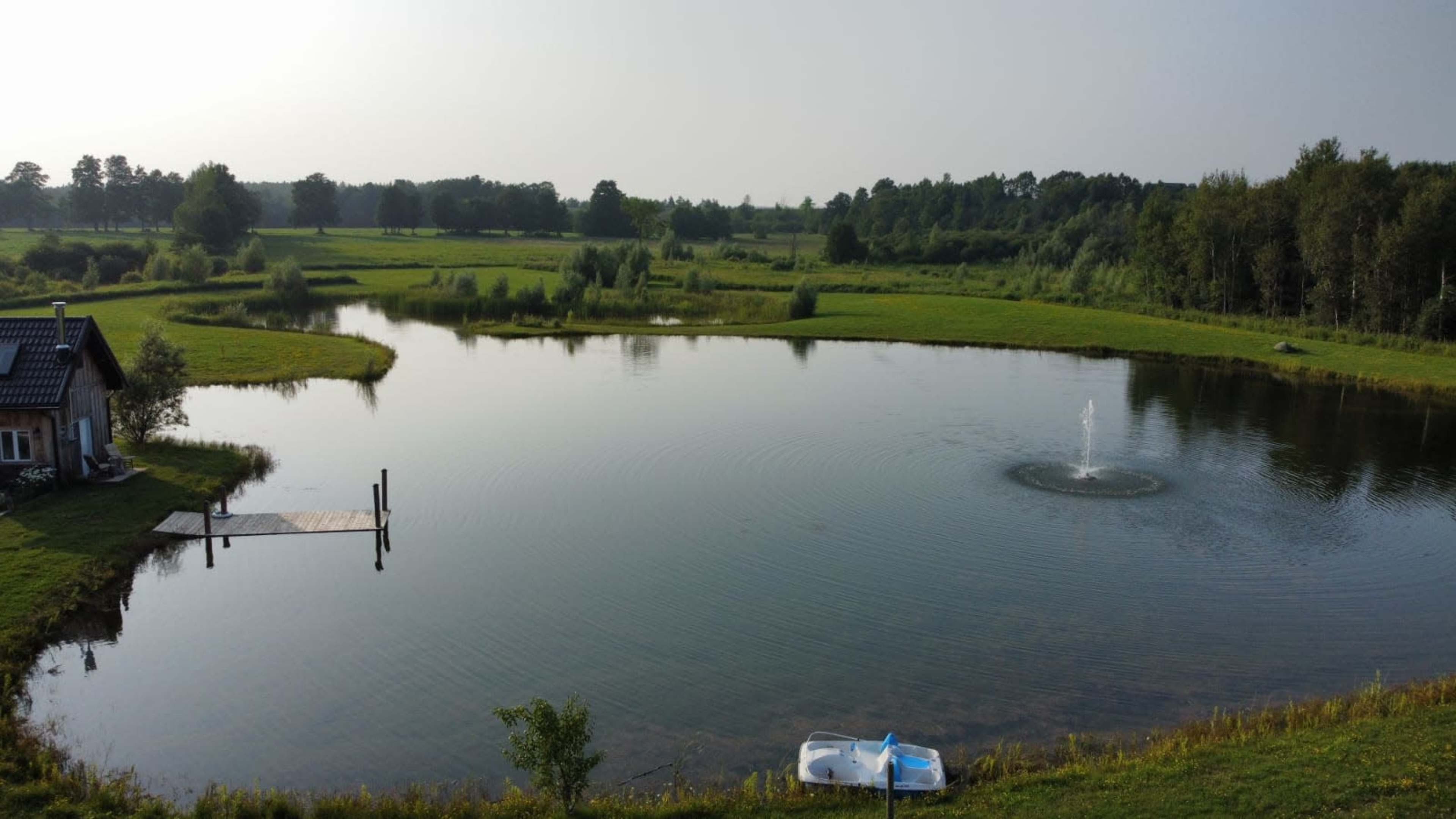A calm pond surrounded by lush green fields and trees, with a dock on the left and a small boat partially visible in the foreground.