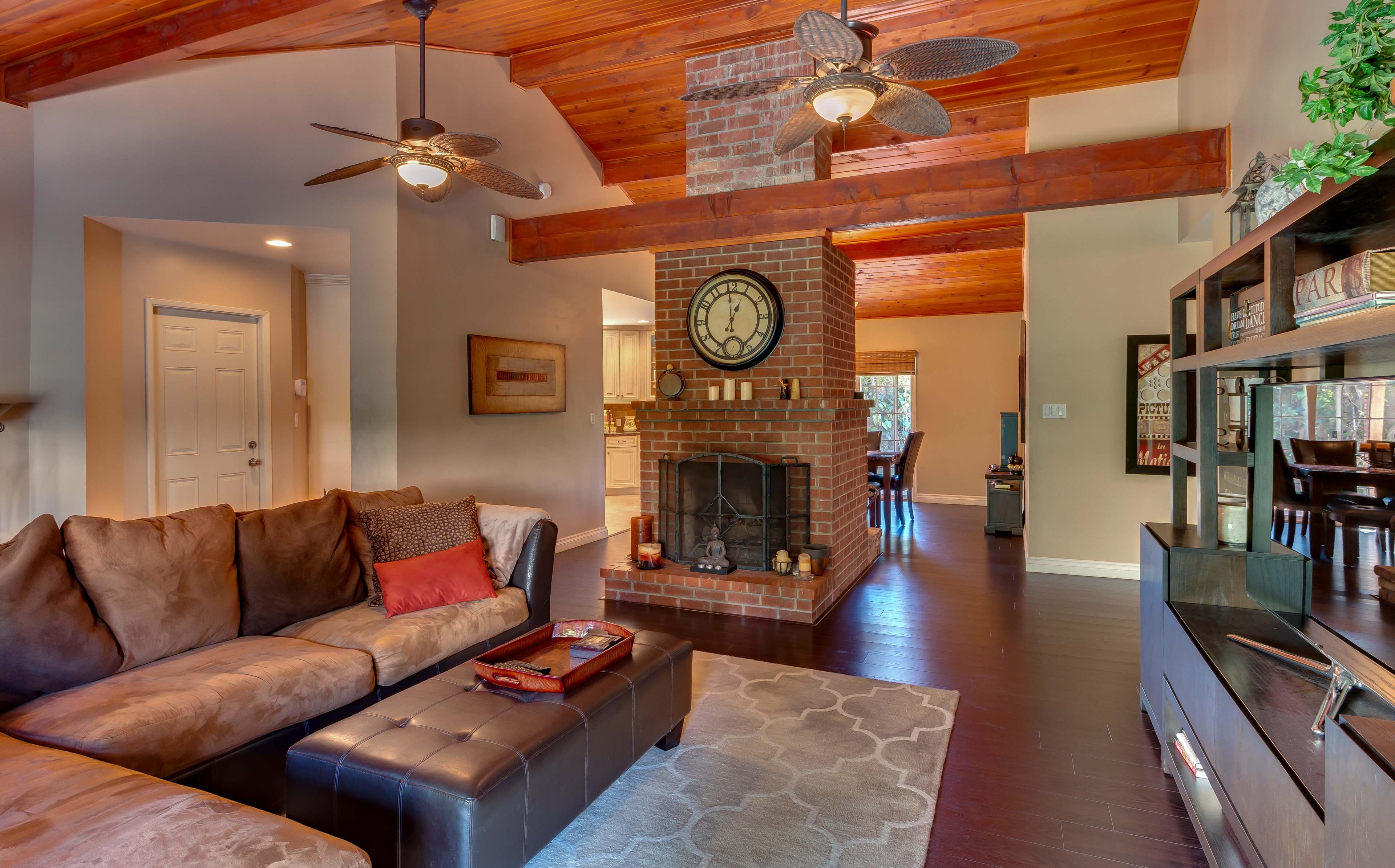 The living room features a brown sectional sofa, a brick fireplace with a round clock above it, and wooden beams on the ceiling.