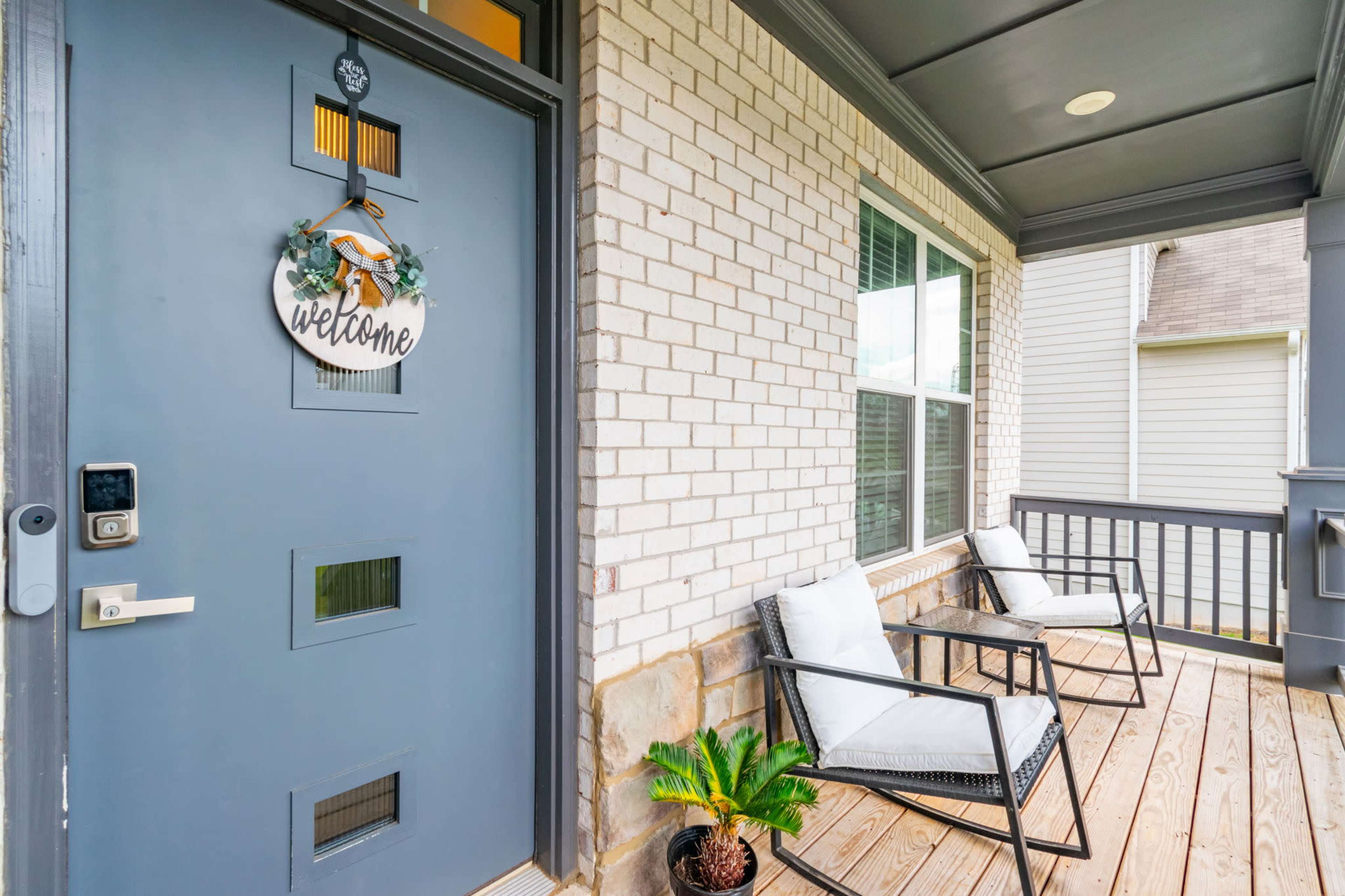 A front porch with a blue door, a welcome sign, two chairs, and a small potted plant.
