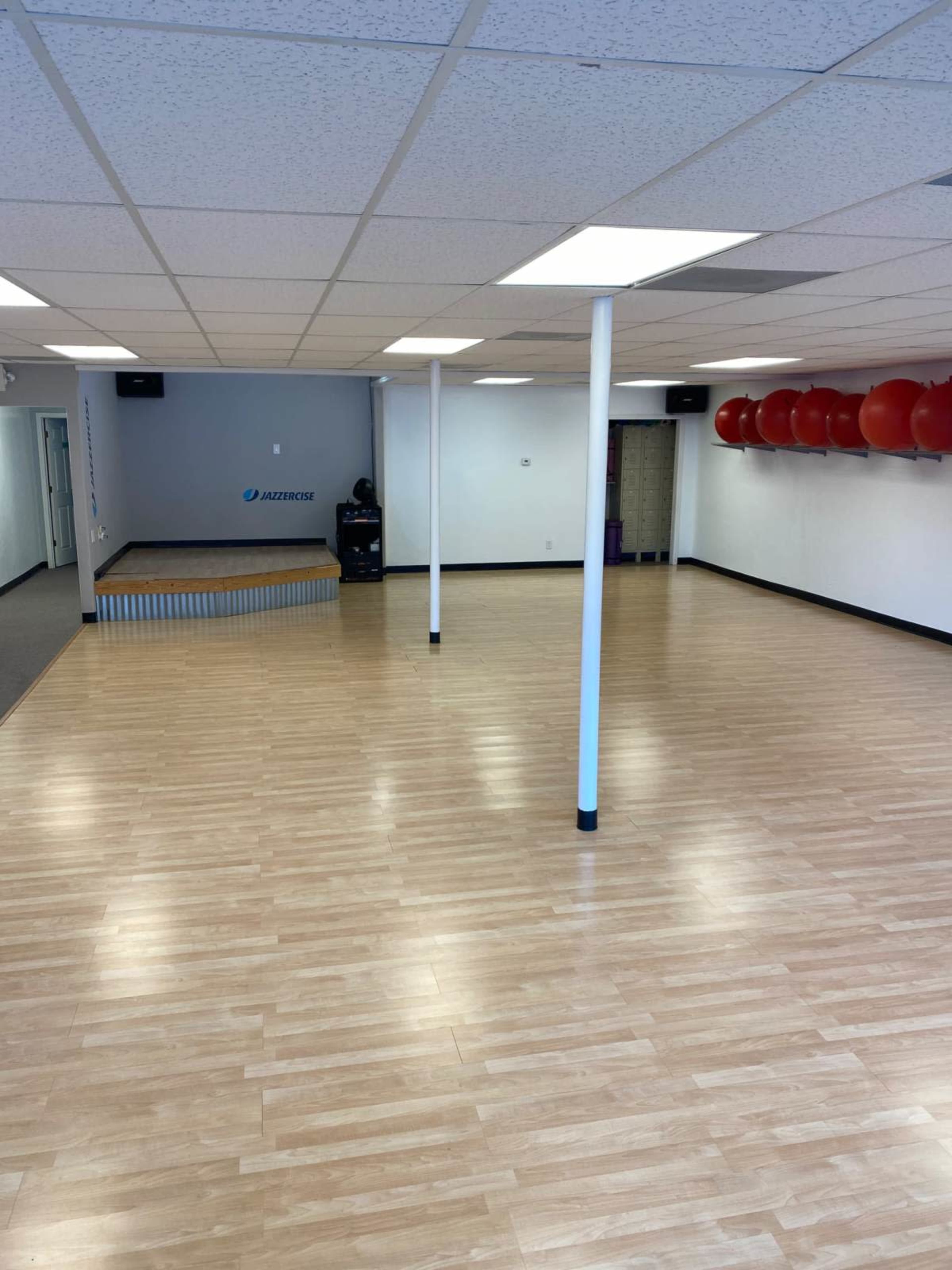 An empty fitness studio with a wooden floor, white walls, and exercise balls in red on a shelf.