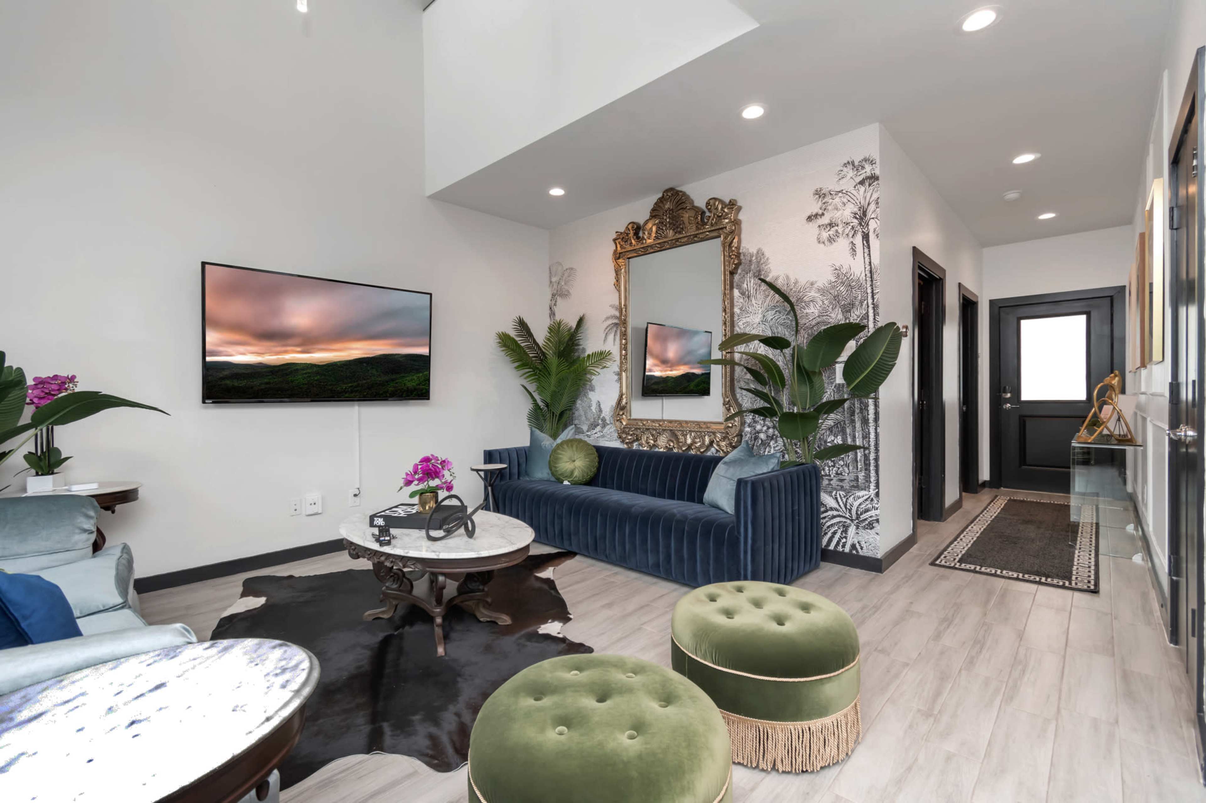 A modern living room with a blue velvet sofa, green ottomans, a large mirror, and a television displaying a landscape, surrounded by decorative plants and a patterned wall.