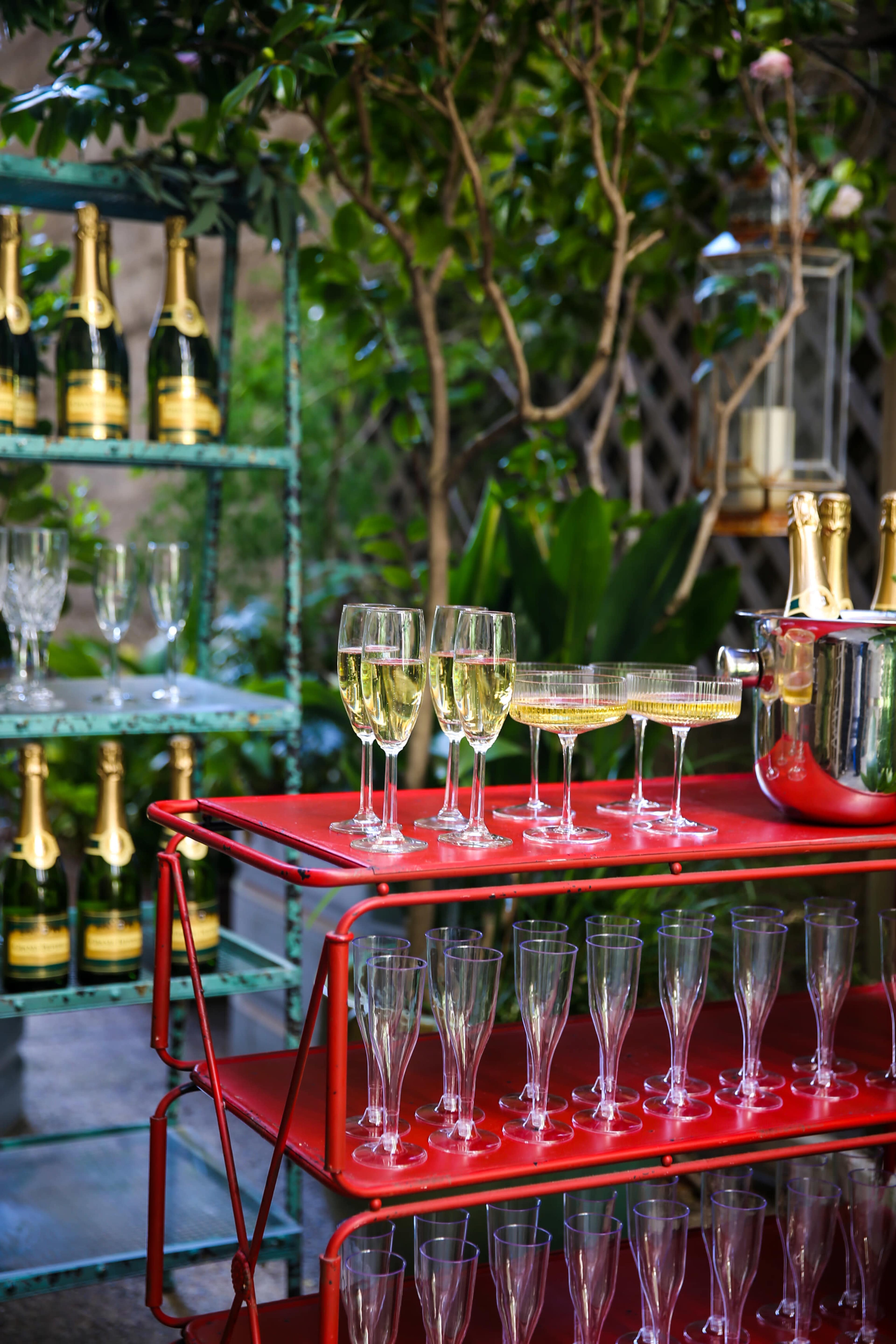 A red metal cart displays several champagne flutes and a bottle bucket alongside a shelf of champagne bottles in a garden setting.