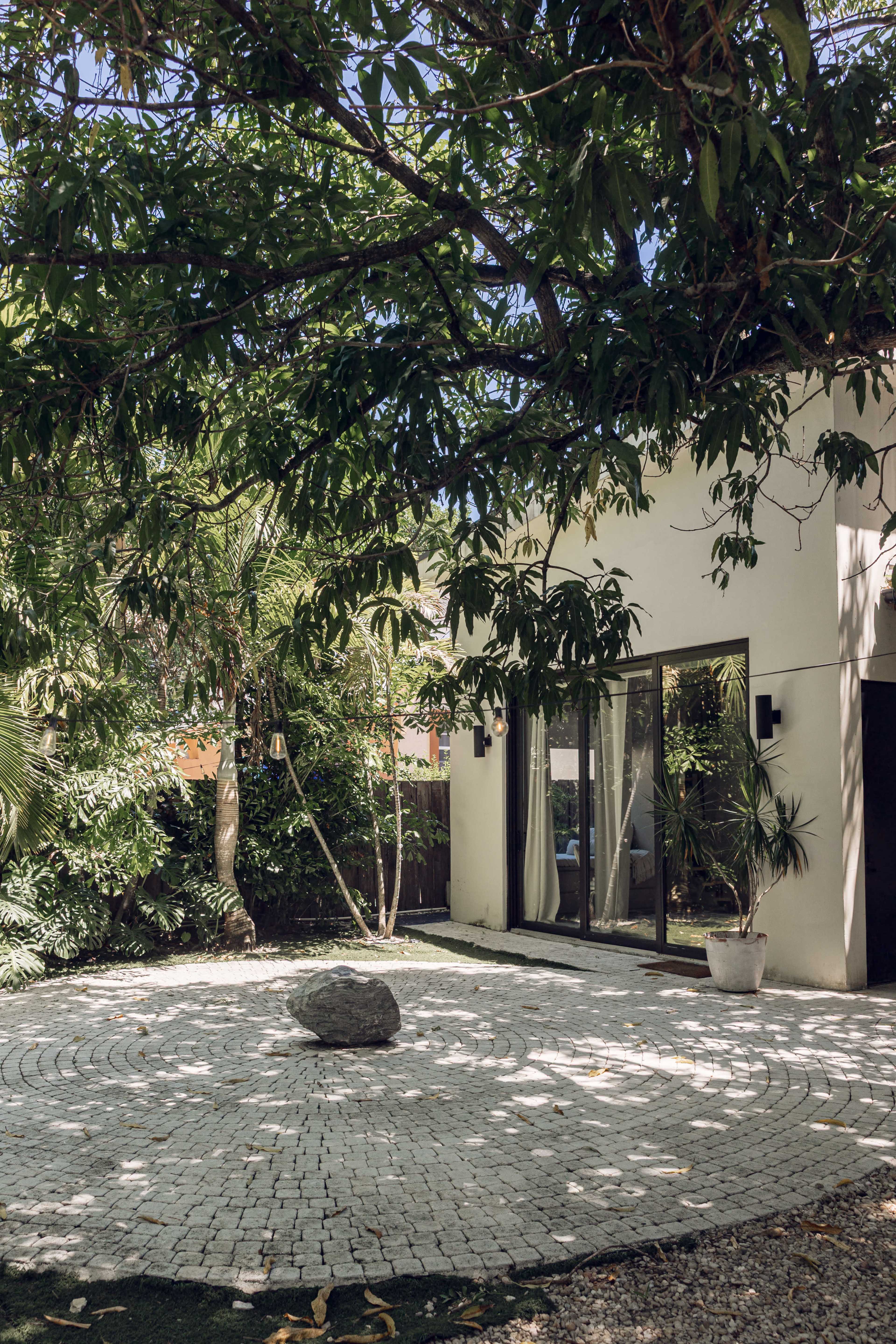 A modern home with large glass windows is set in a landscaped yard featuring a circular stone patio and surrounded by lush greenery.