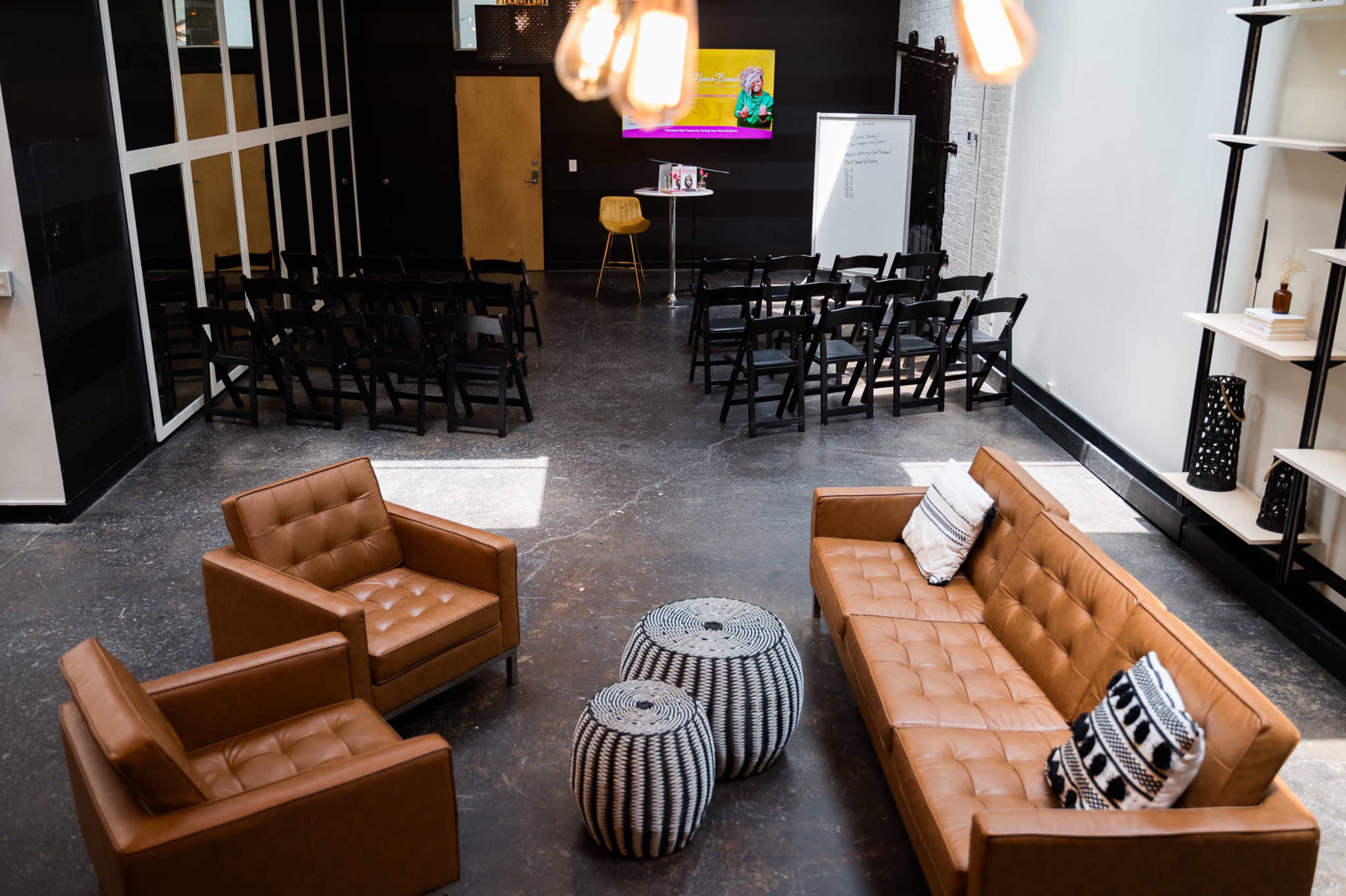 A modern gathering space with brown leather chairs, circular patterned poufs, and rows of folding chairs facing a presentation screen.
