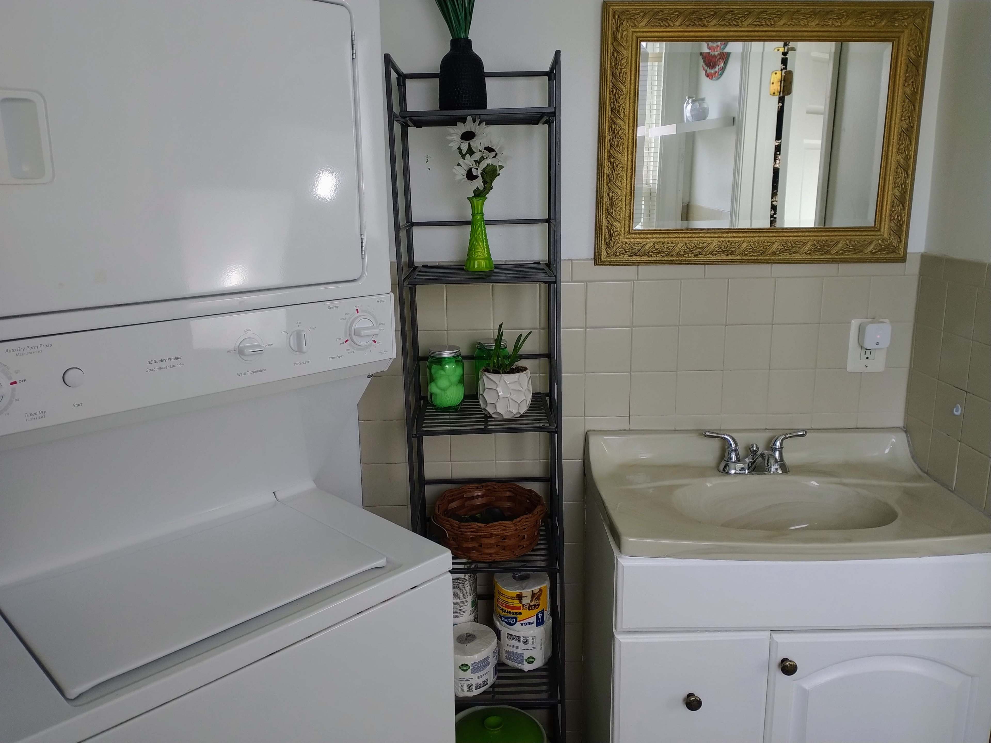 A laundry room with a stacked washer and dryer on the left, a small shelf with decorative items on the right, and a sink below a mirror.
