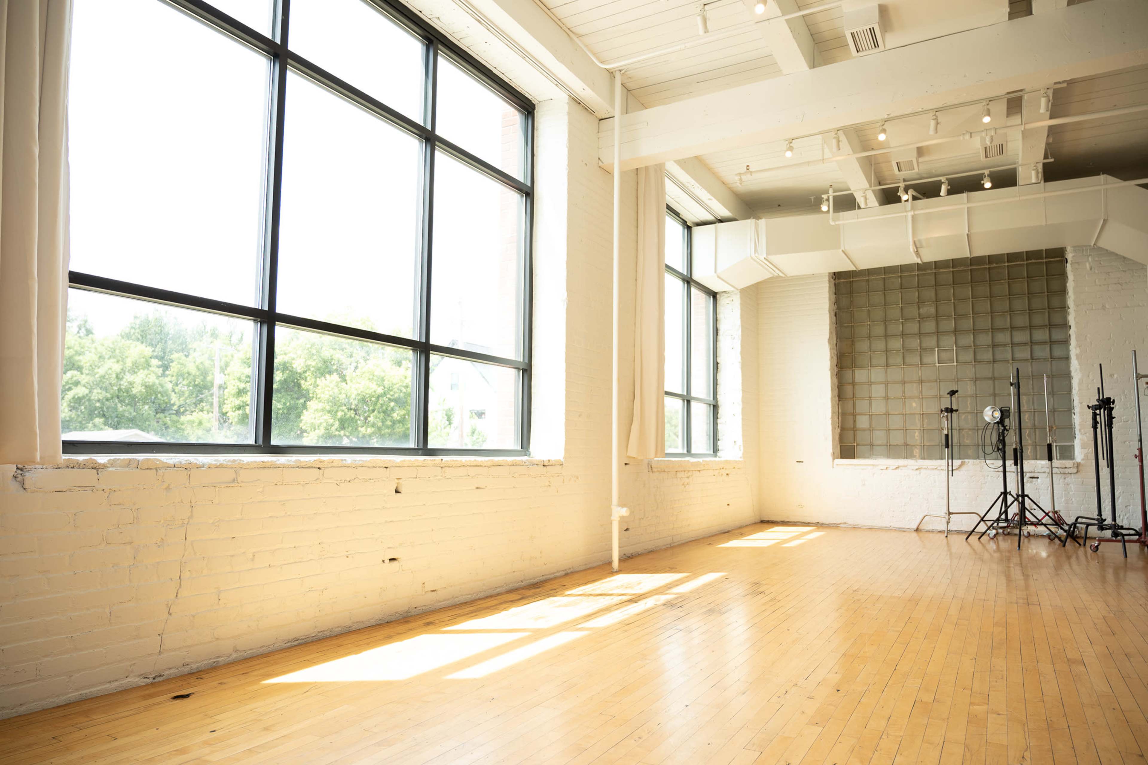 A spacious room with large windows letting in natural light, featuring hardwood floors and an empty wall with studio equipment in the background.