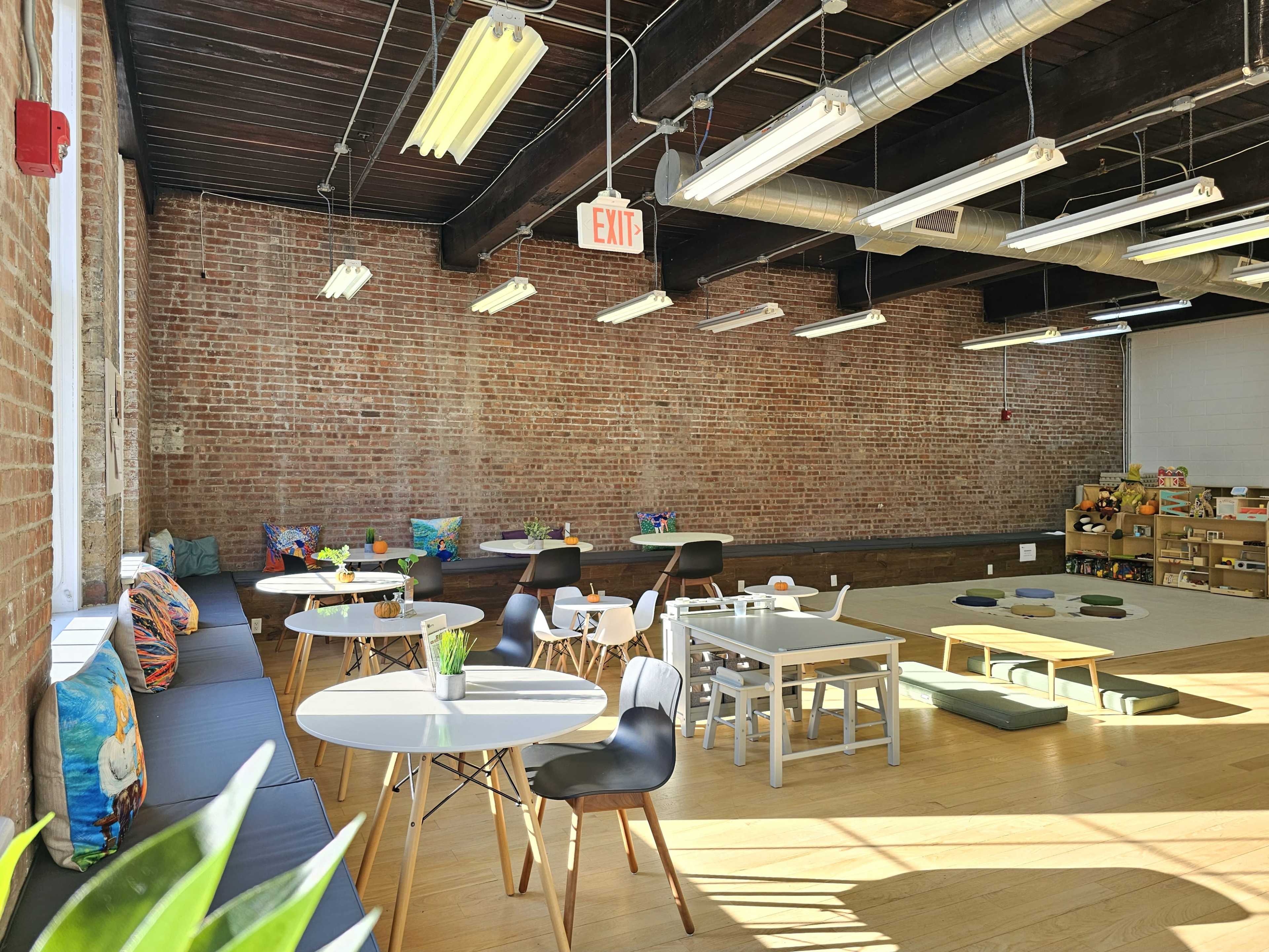 The image shows a spacious, modern room with exposed brick walls, several tables and chairs, and a variety of colorful cushions scattered around.