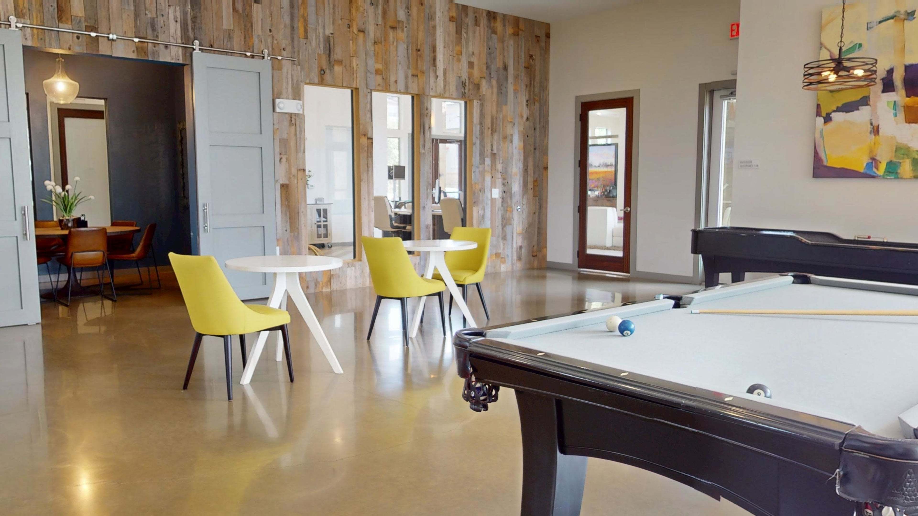 The image features a modern interior space with a pool table in the foreground, a wooden accent wall, and seating areas that include round tables with yellow chairs.