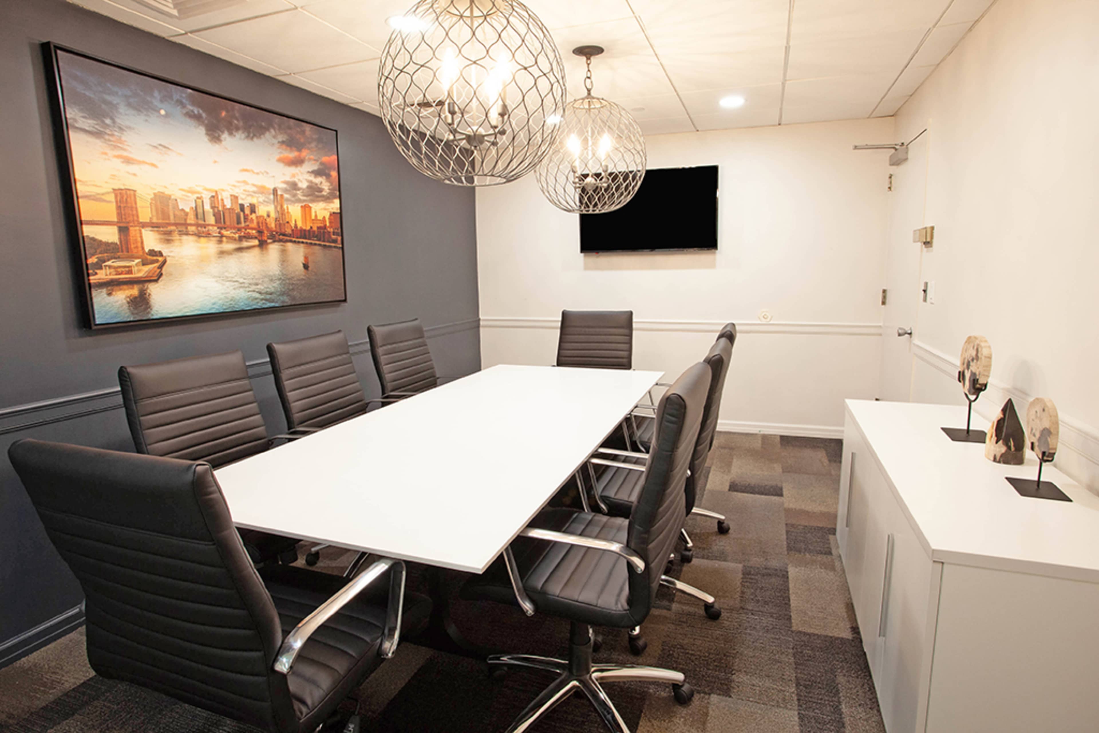 The image shows a modern conference room featuring a long white table surrounded by black office chairs, a large cityscape artwork on the wall, and pendant lighting.
