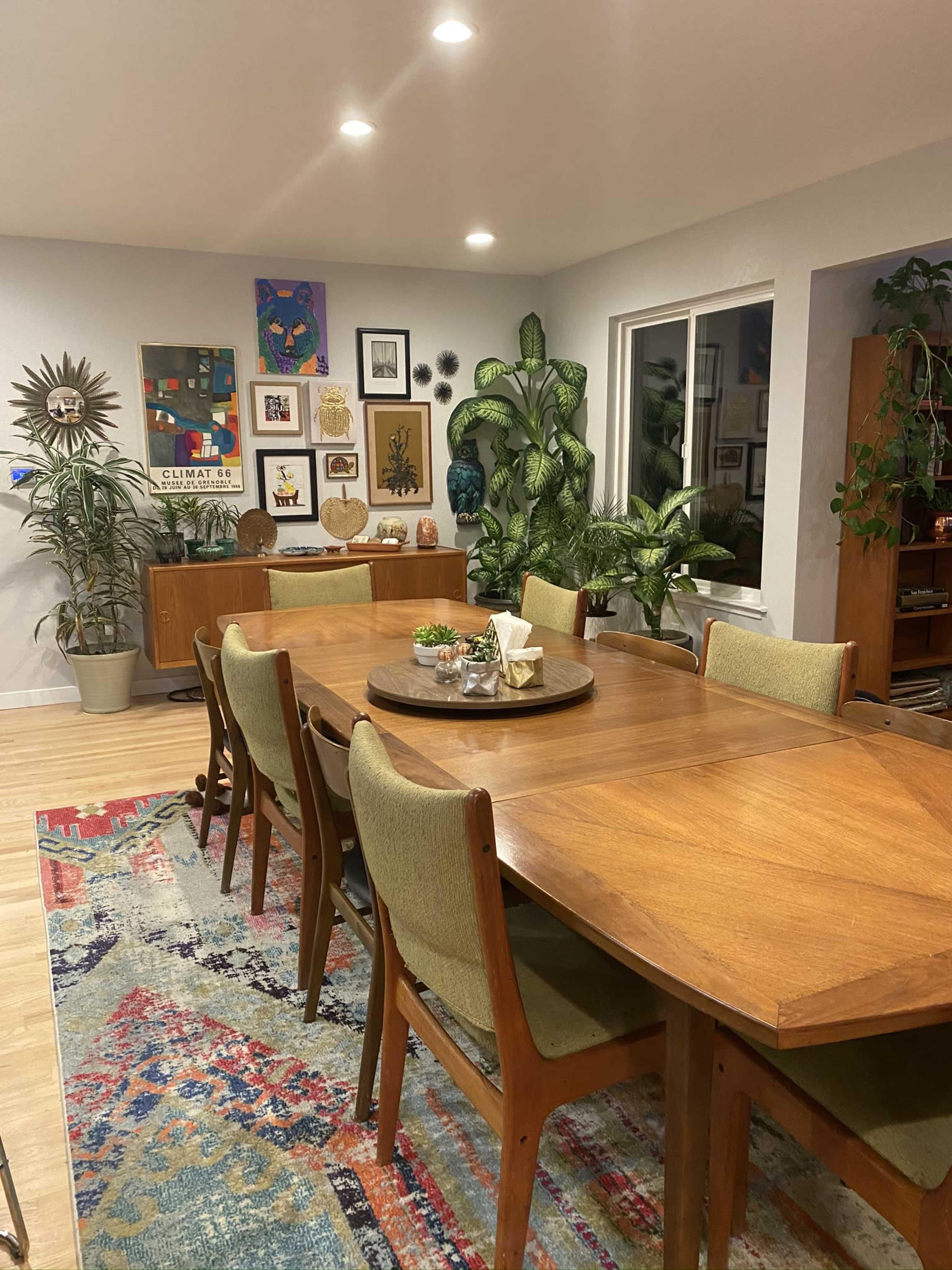 A wooden dining table surrounded by green upholstered chairs is set in a brightly lit room decorated with various artworks and indoor plants.