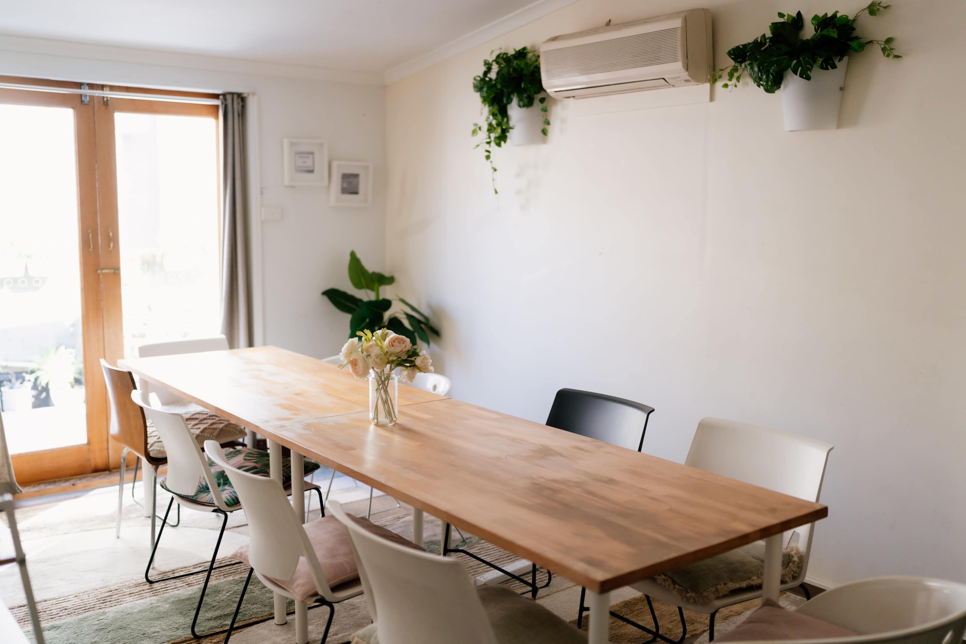 The image shows a dining area featuring a long wooden table with a vase of flowers, surrounded by chairs, and a wall with a plant and air conditioning unit.