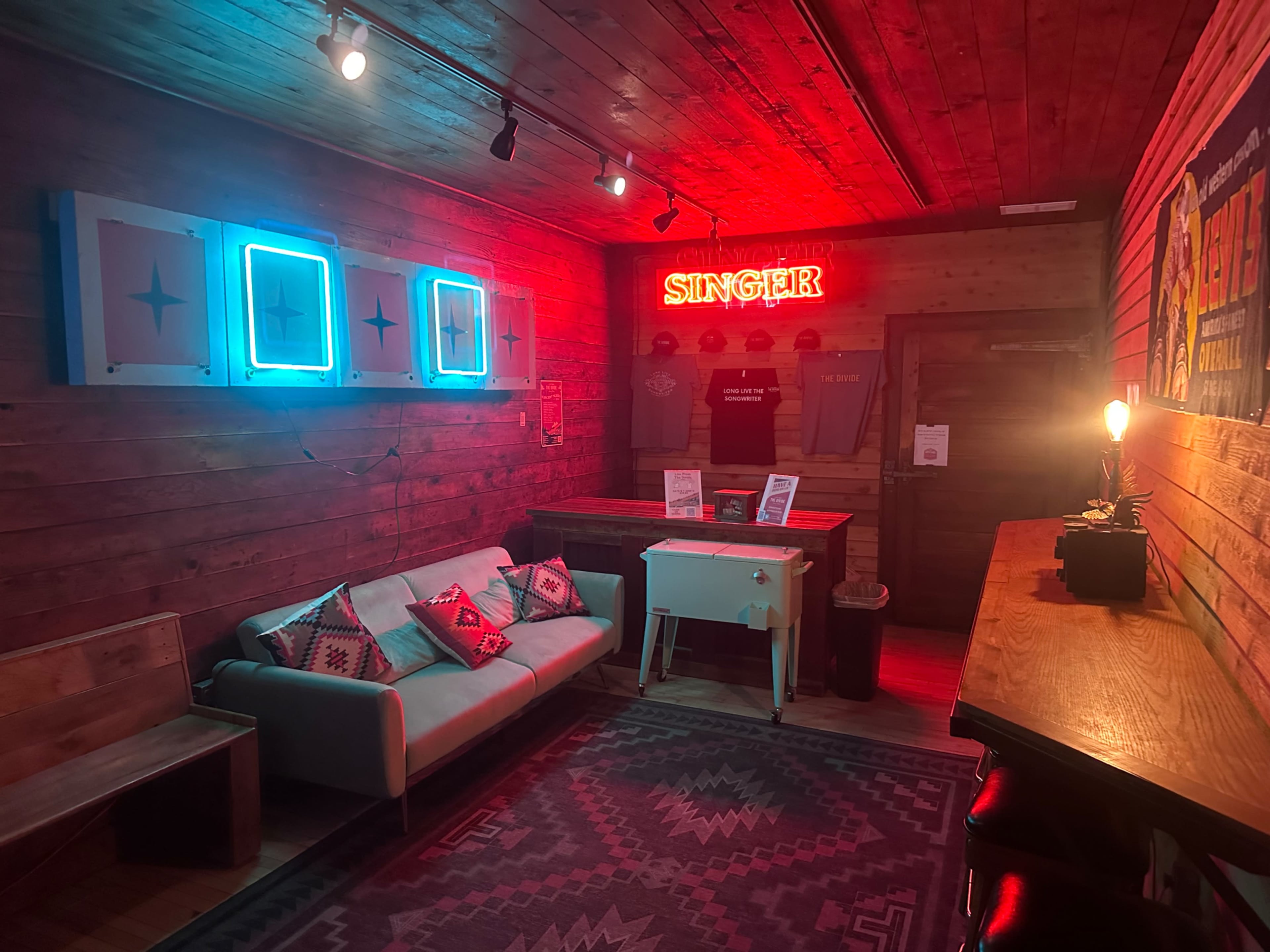 The interior features a cozy room with wooden walls, a couch, a red neon sign reading "SINGER," and a small table amidst contrasting decorative elements.