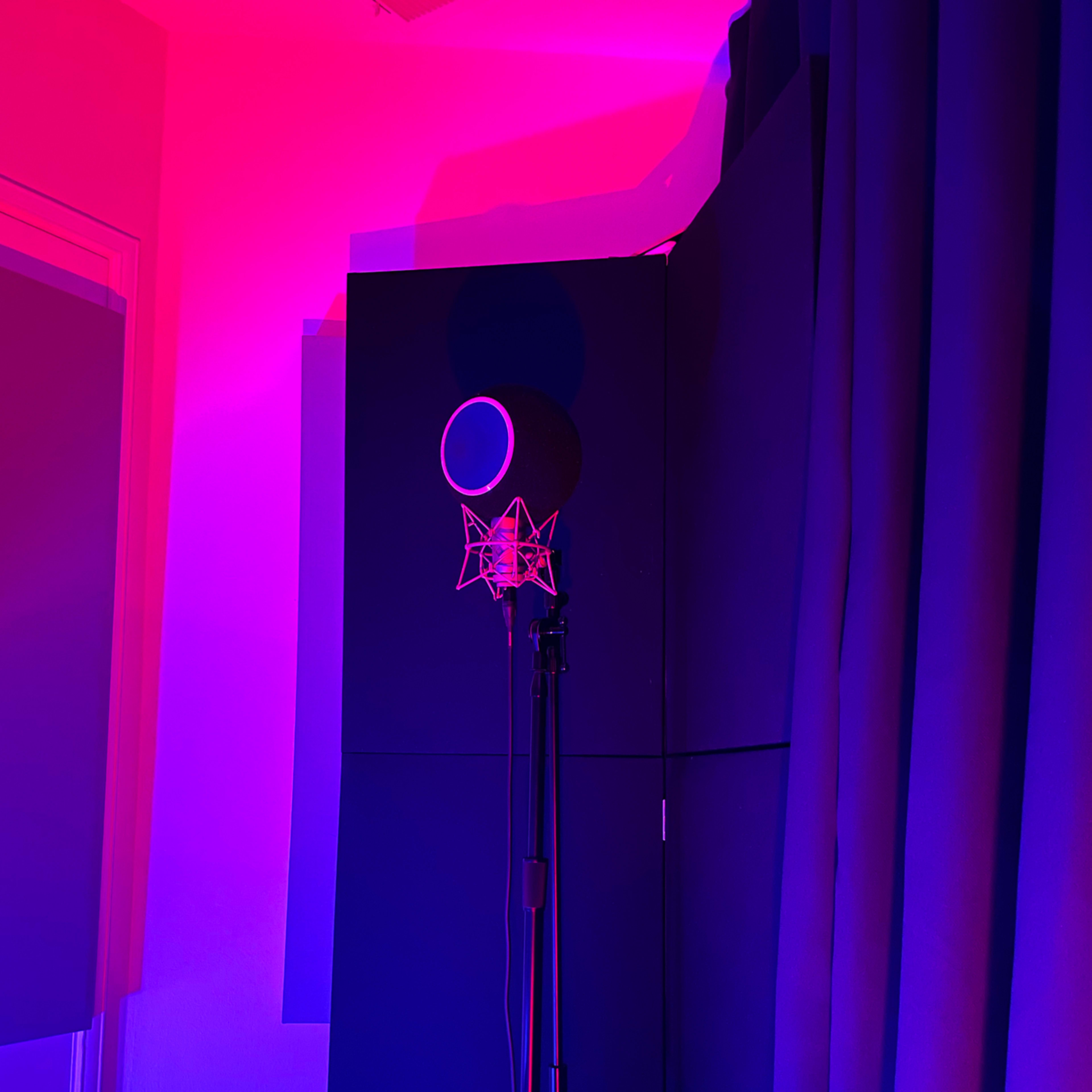 A microphone on a stand is positioned in front of dark soundproofing panels illuminated by pink and purple lighting.
