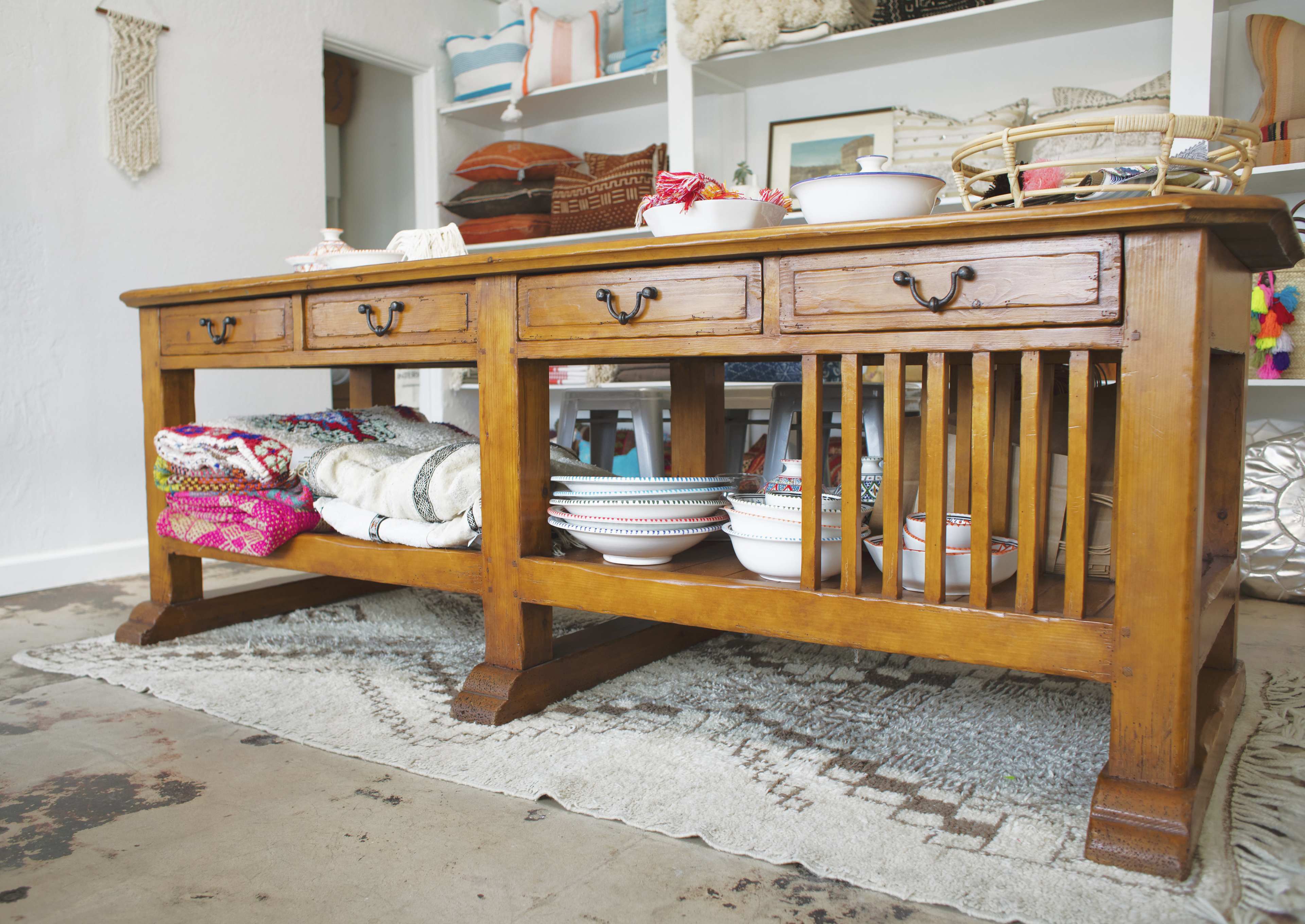A wooden console table with drawers and open shelving displays various decorative bowls and textiles in a bright, airy room.
