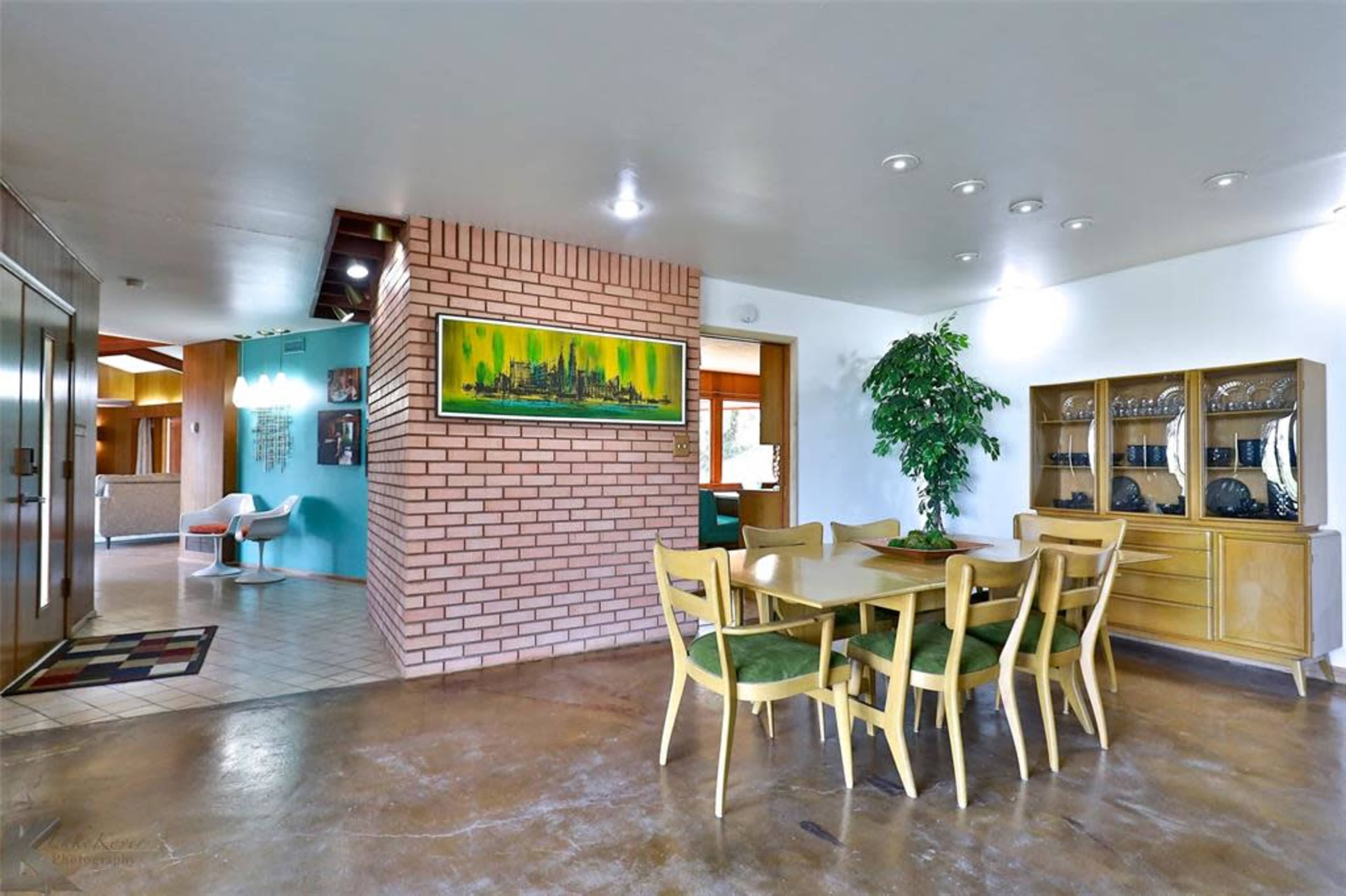 The image shows a dining area with a green table and yellow chairs, situated next to a brick wall and a glass display cabinet.