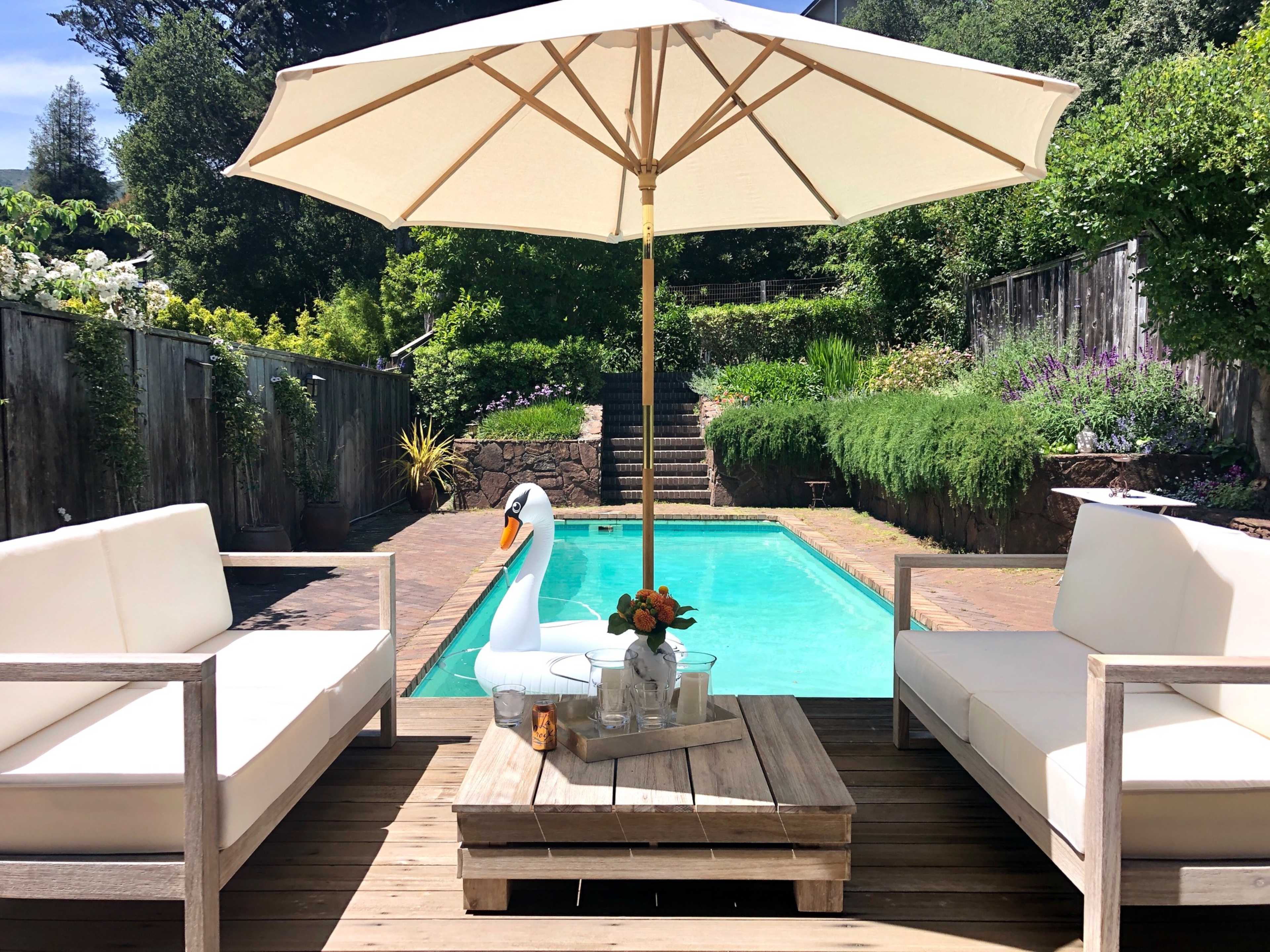 A patio area features two white couches and a wooden table beside a swimming pool, shaded by a large umbrella.