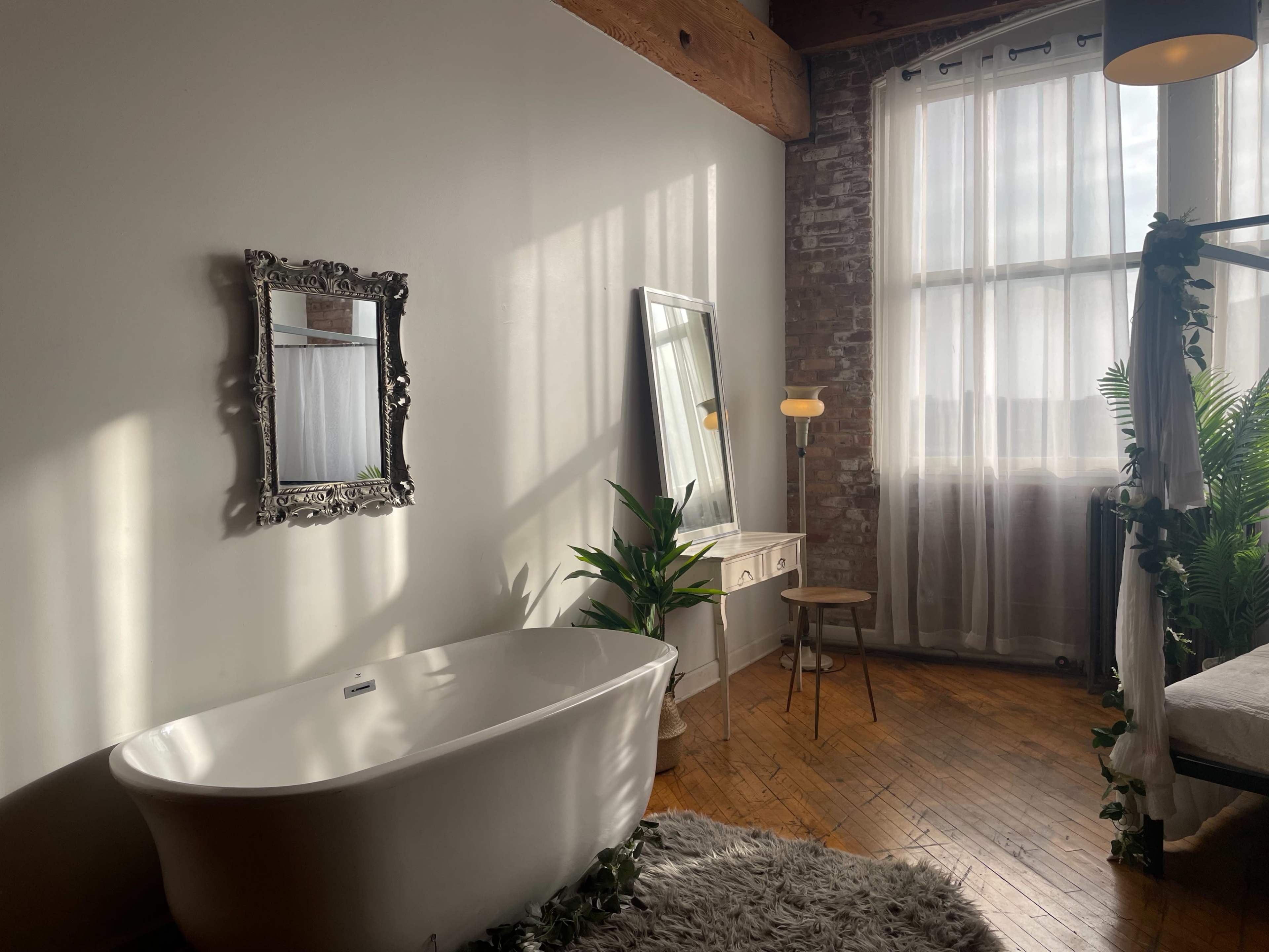 A spacious bathroom featuring a freestanding white bathtub, a large mirror, a wooden chair, and a cozy interior with natural light illuminating the space.