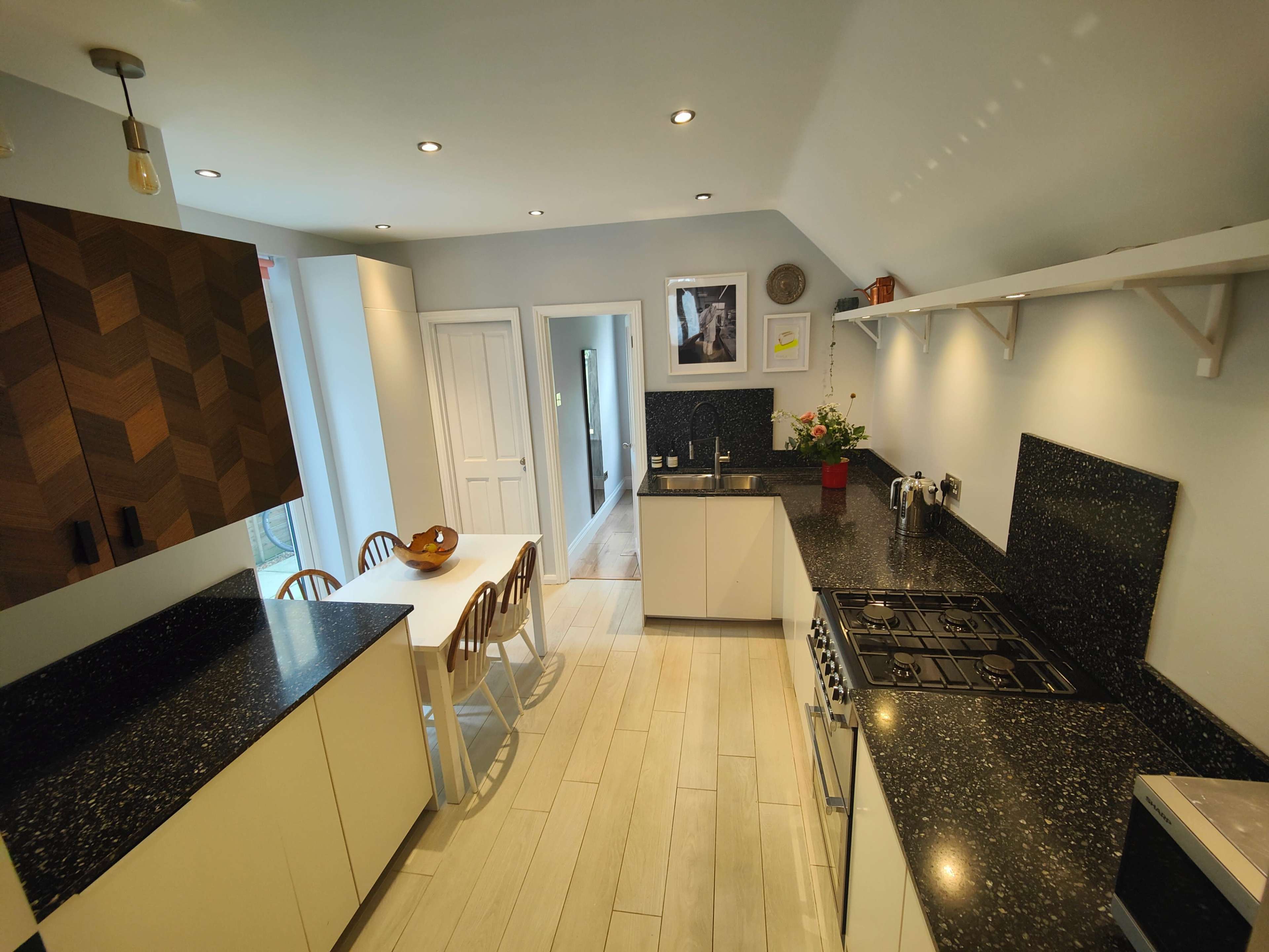 A modern kitchen with dark countertops, light-colored cabinetry, and a dining table with wooden chairs, leading to a hallway.