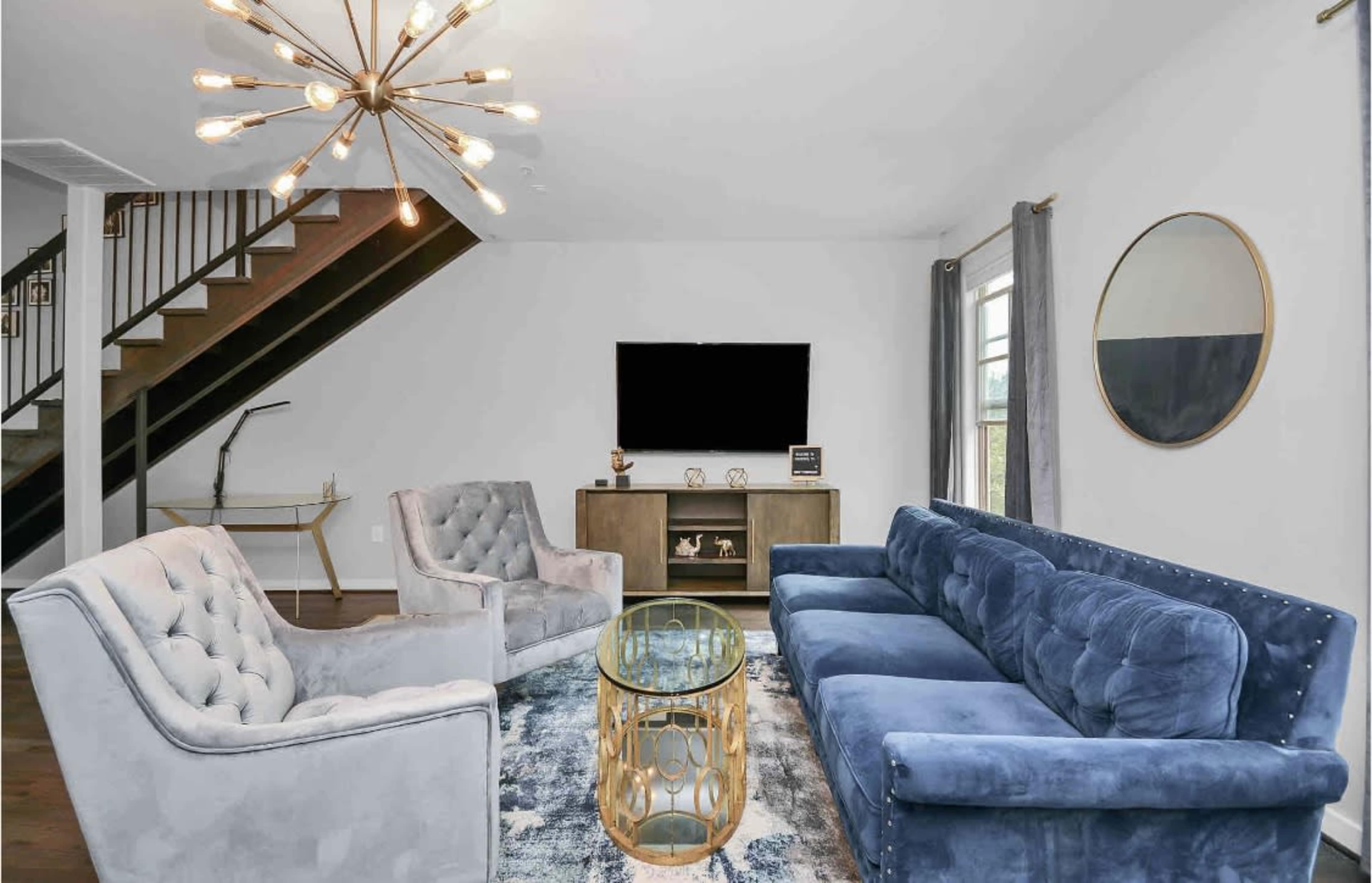 The image shows a modern living room featuring a blue velvet sofa, two gray armchairs, a glass coffee table, and a television mounted on the wall, with a staircase in the background.