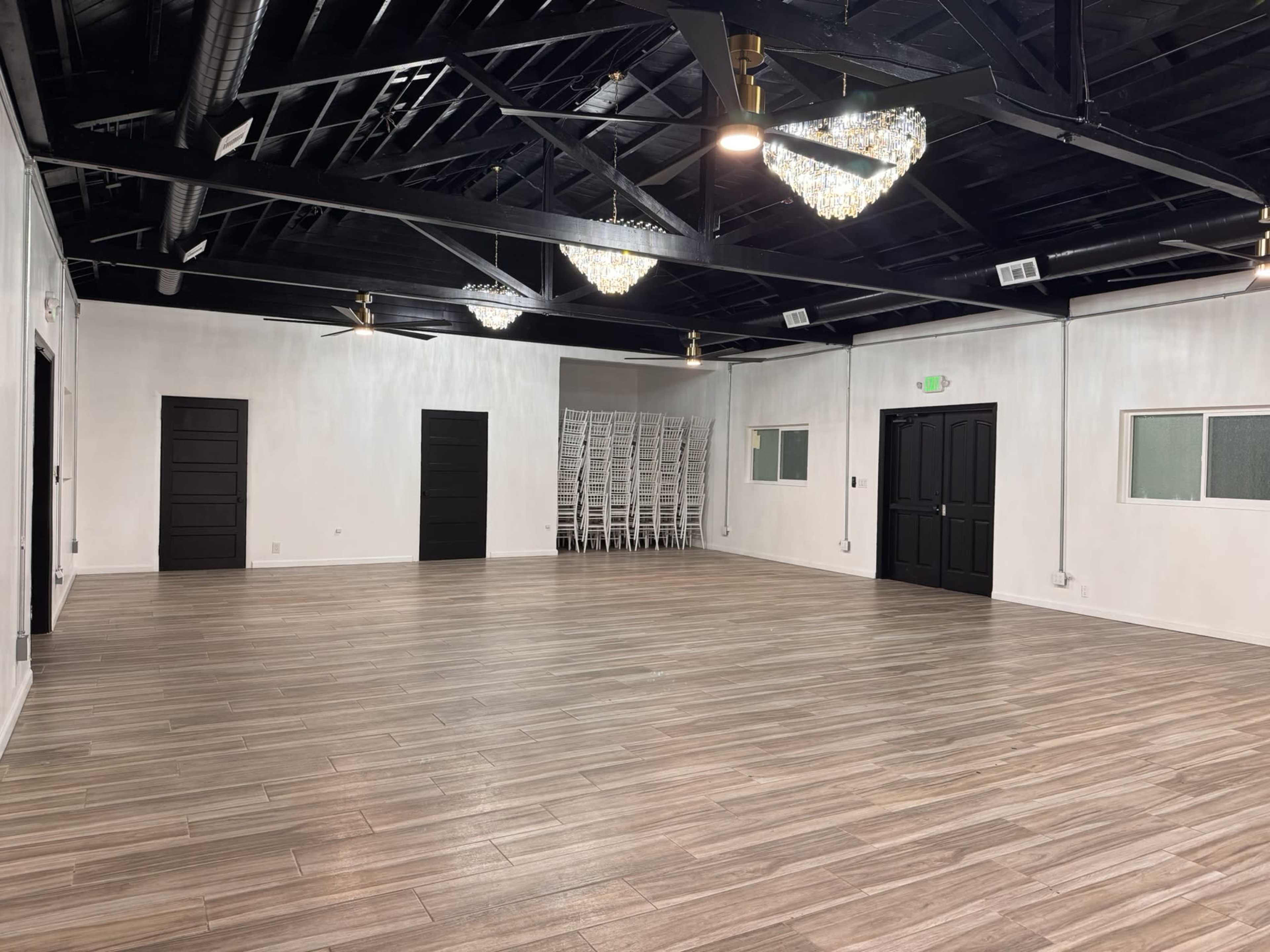 The image shows a spacious, well-lit room with a sleek wooden floor, black ceiling beams, and two ornate chandeliers.