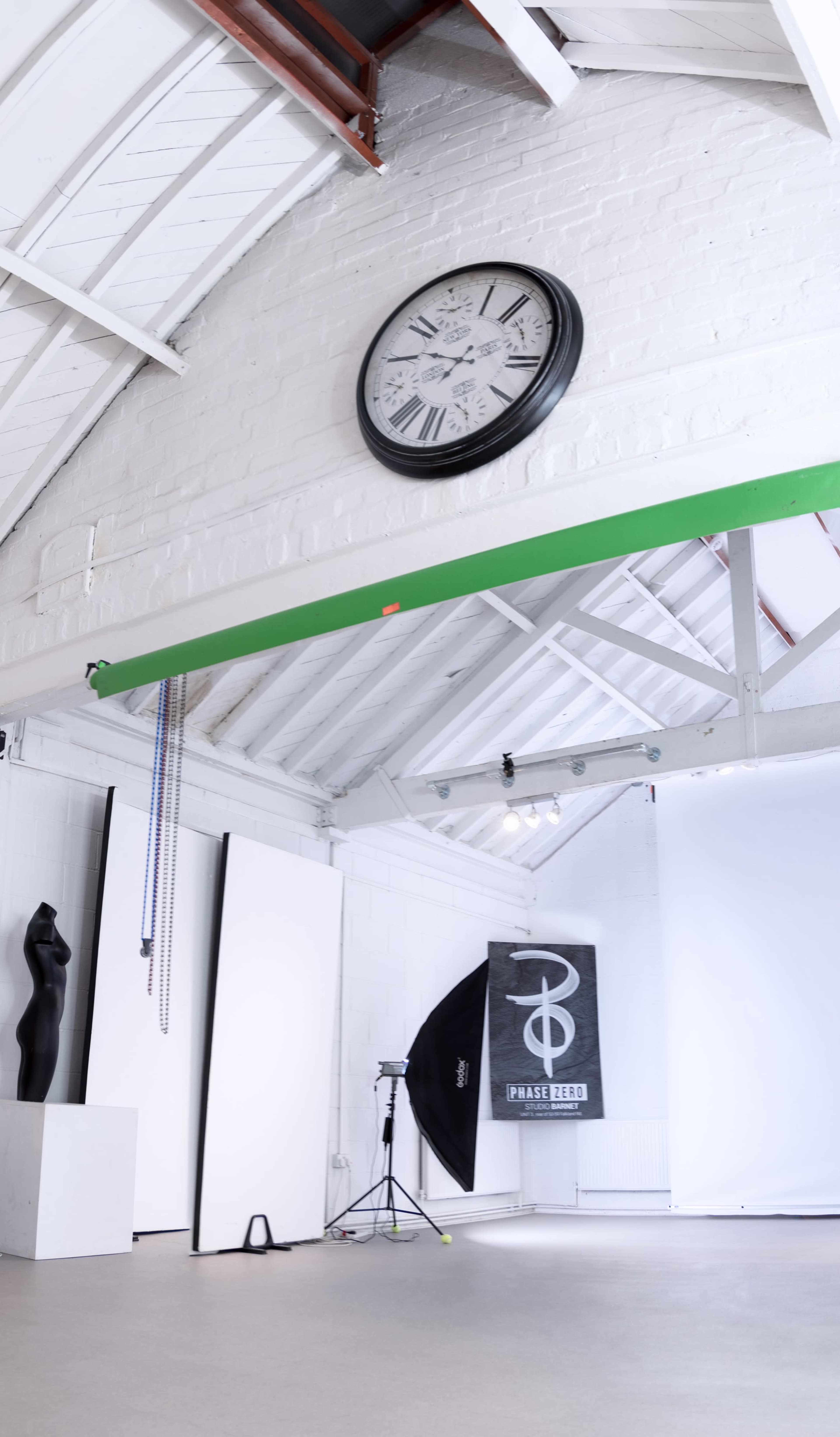 A spacious photography studio with a large clock on the wall, a green beam overhead, and various photography equipment including a softbox and backdrop.