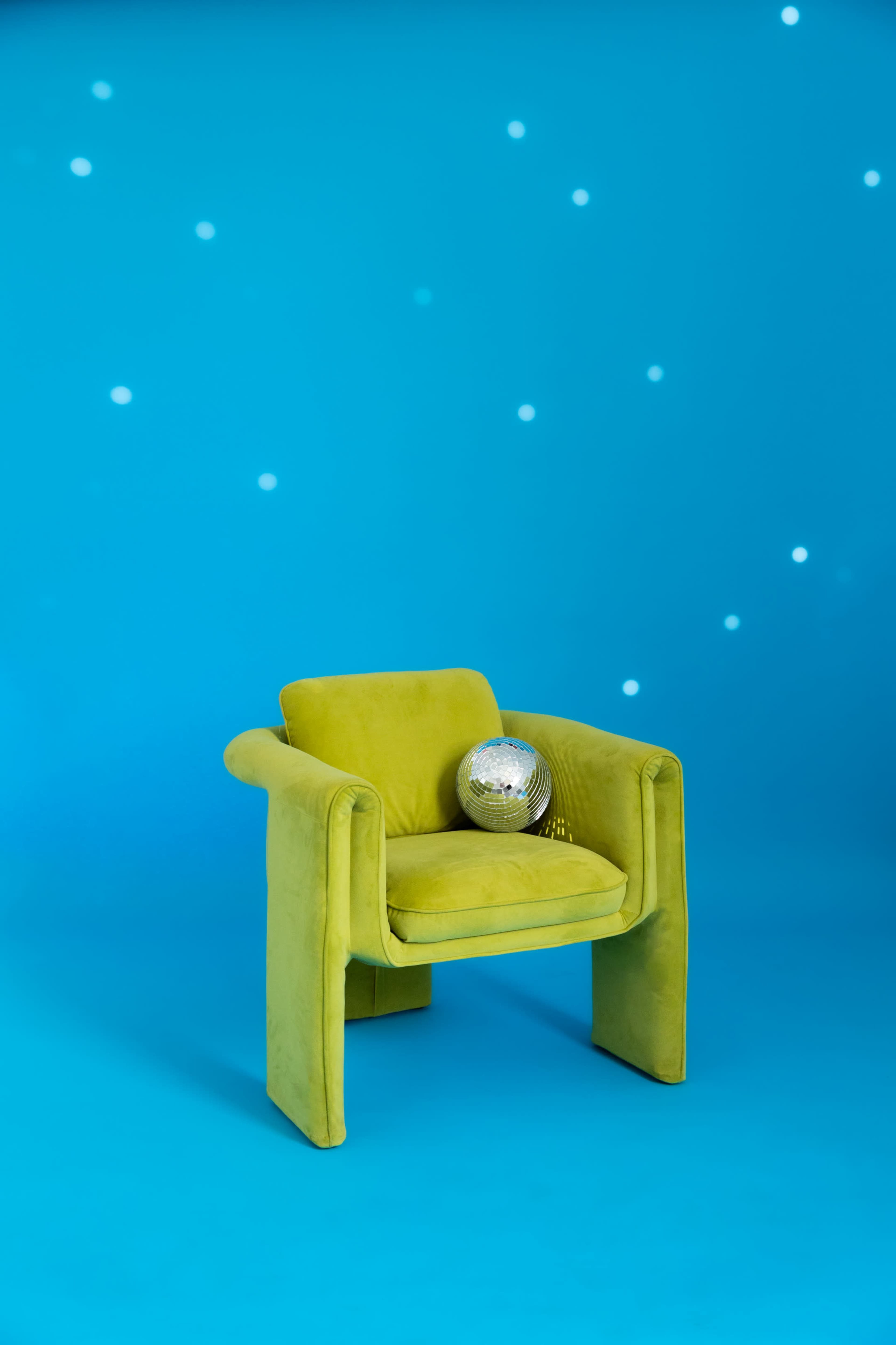 The image features a bright yellow chair with a minimalist design and a disco ball placed on the seat, set against a blue background with scattered light spots.