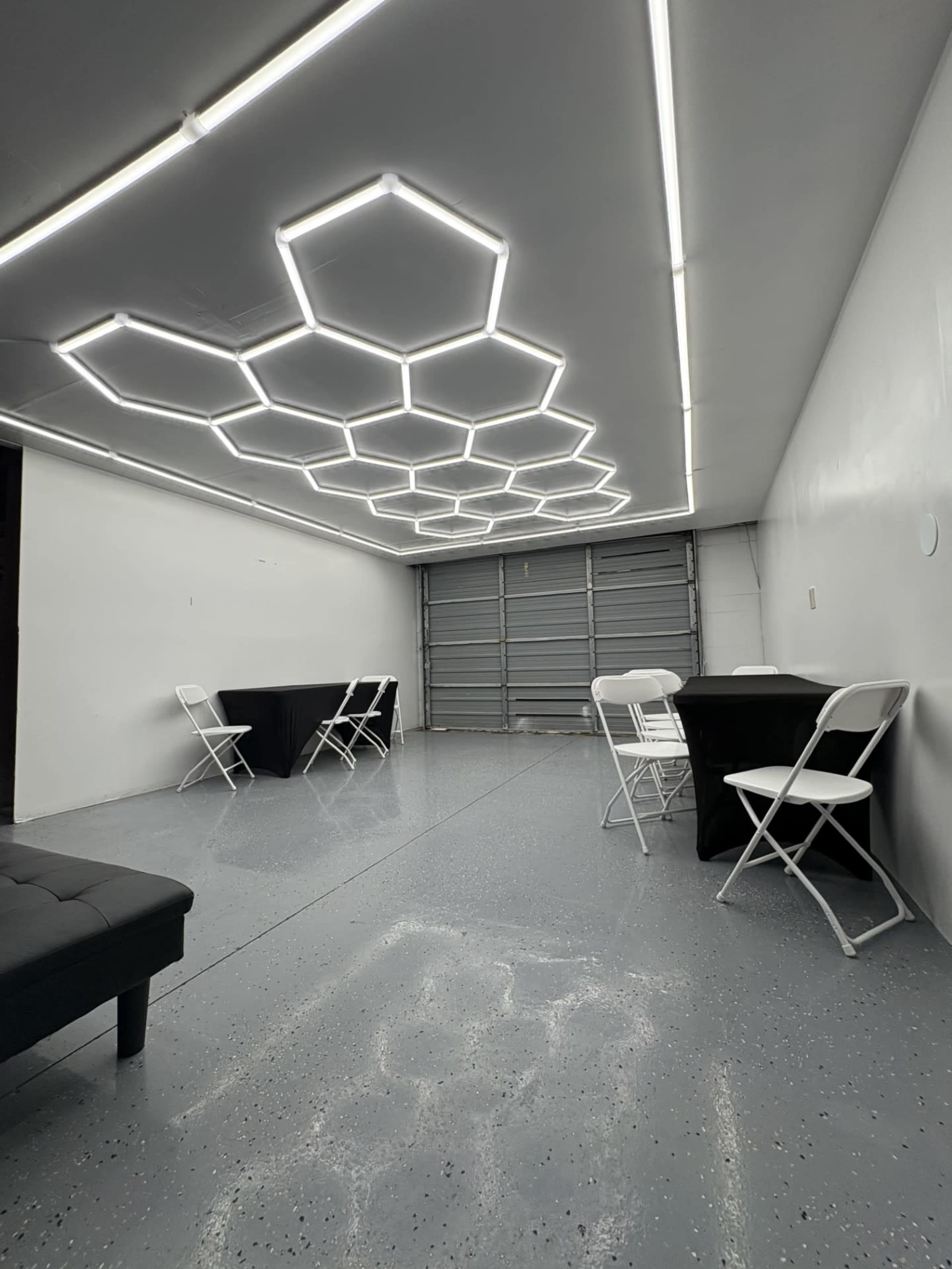 The image shows a modern, minimalist room with a unique hexagonal light fixture on the ceiling and several black tables surrounded by white chairs.