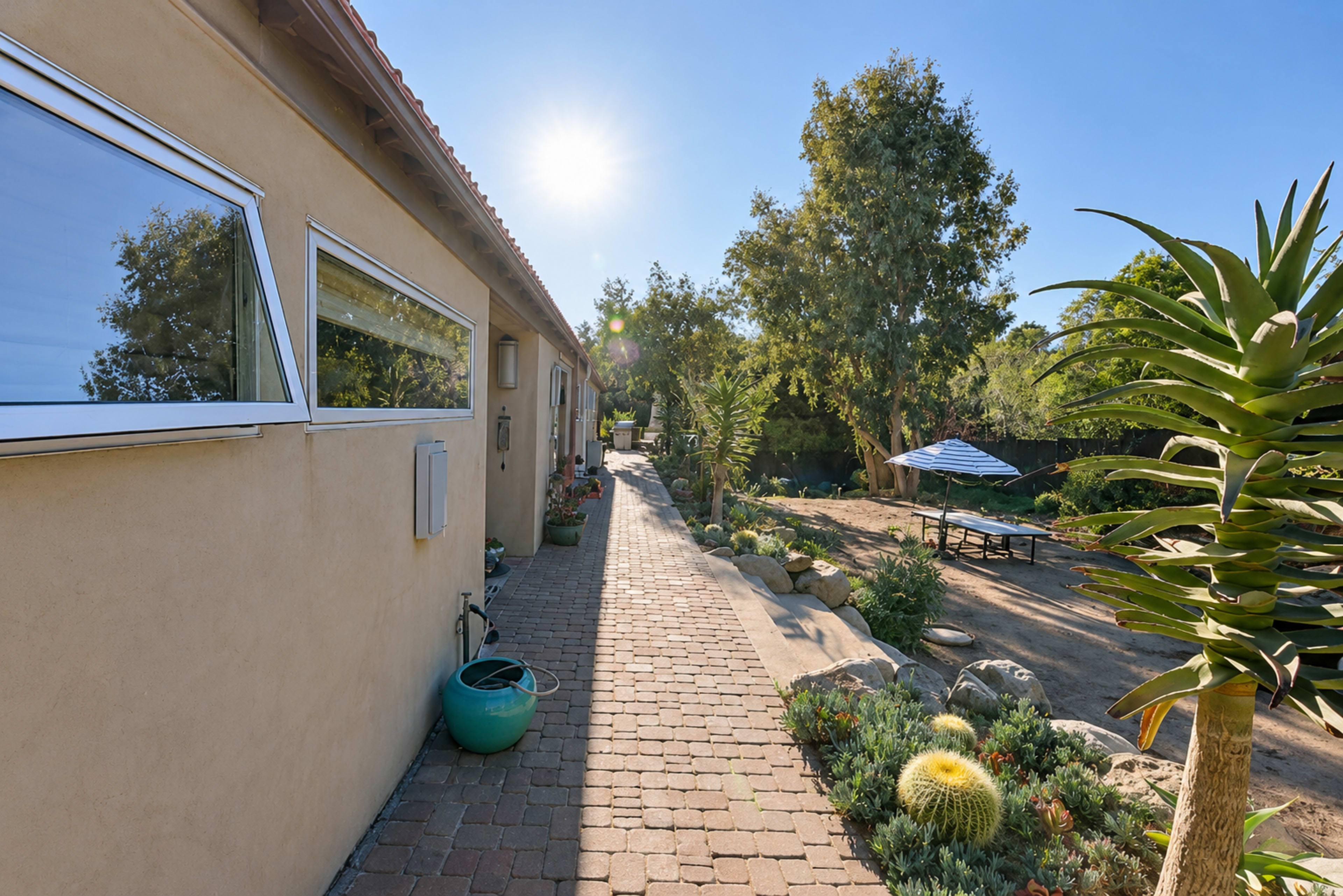 A sunlit pathway lined with plants leads to a patio area under a blue umbrella.