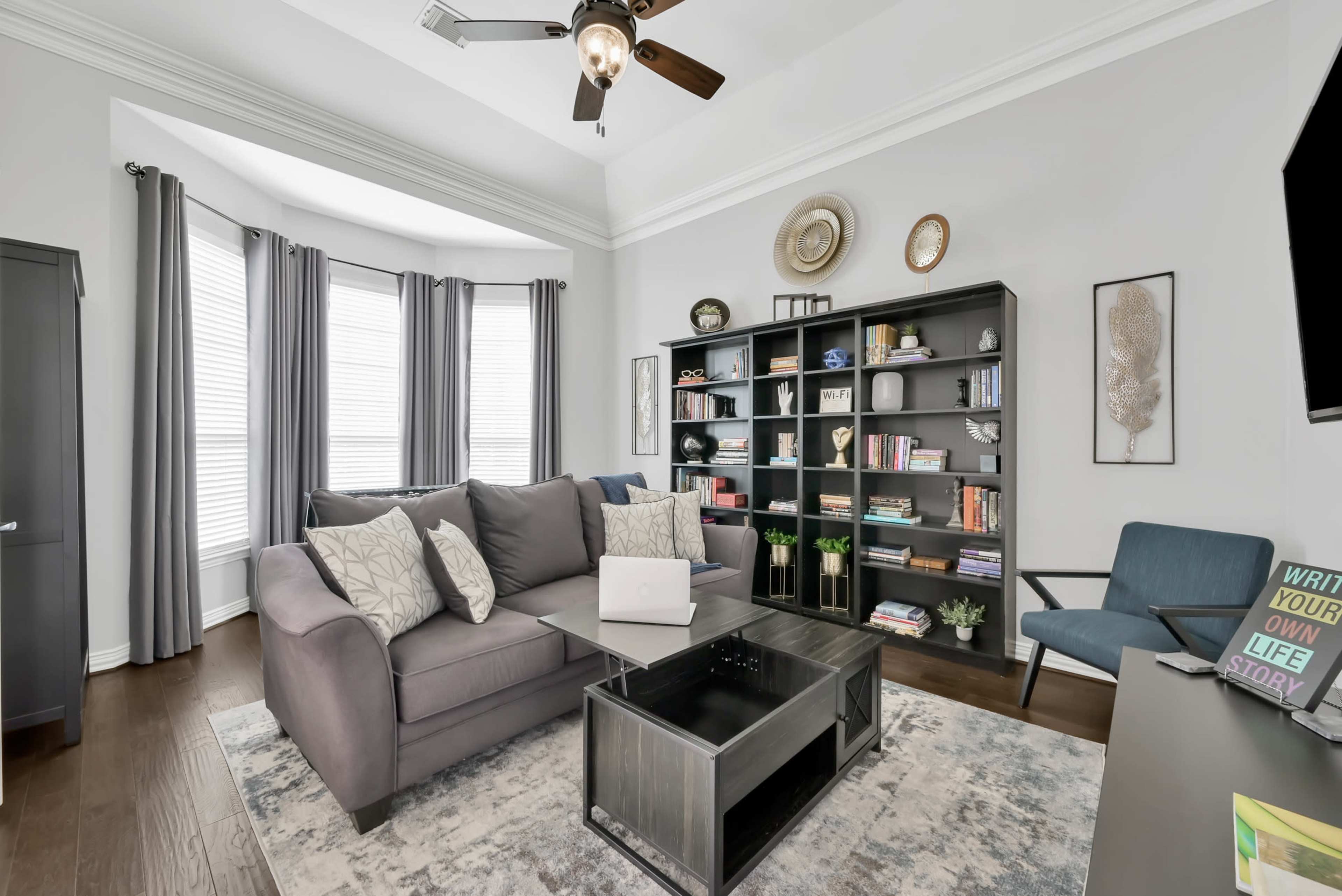 A modern living room features a gray sectional sofa, a black shelving unit filled with books, and large windows with gray curtains.