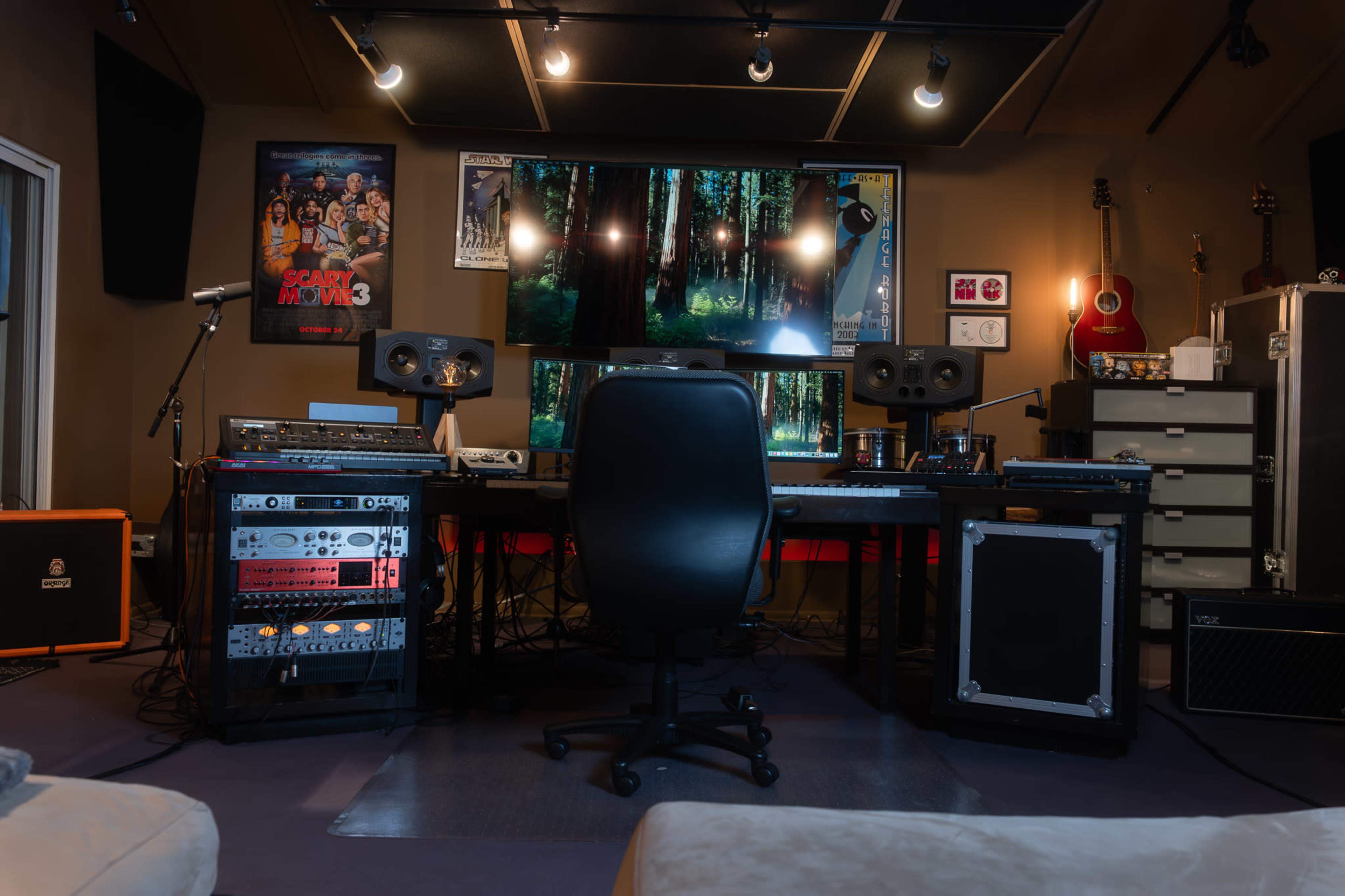 The image shows a modern audio production studio featuring a large desk with audio equipment, a computer display with forest imagery, and musical instruments lining the walls.