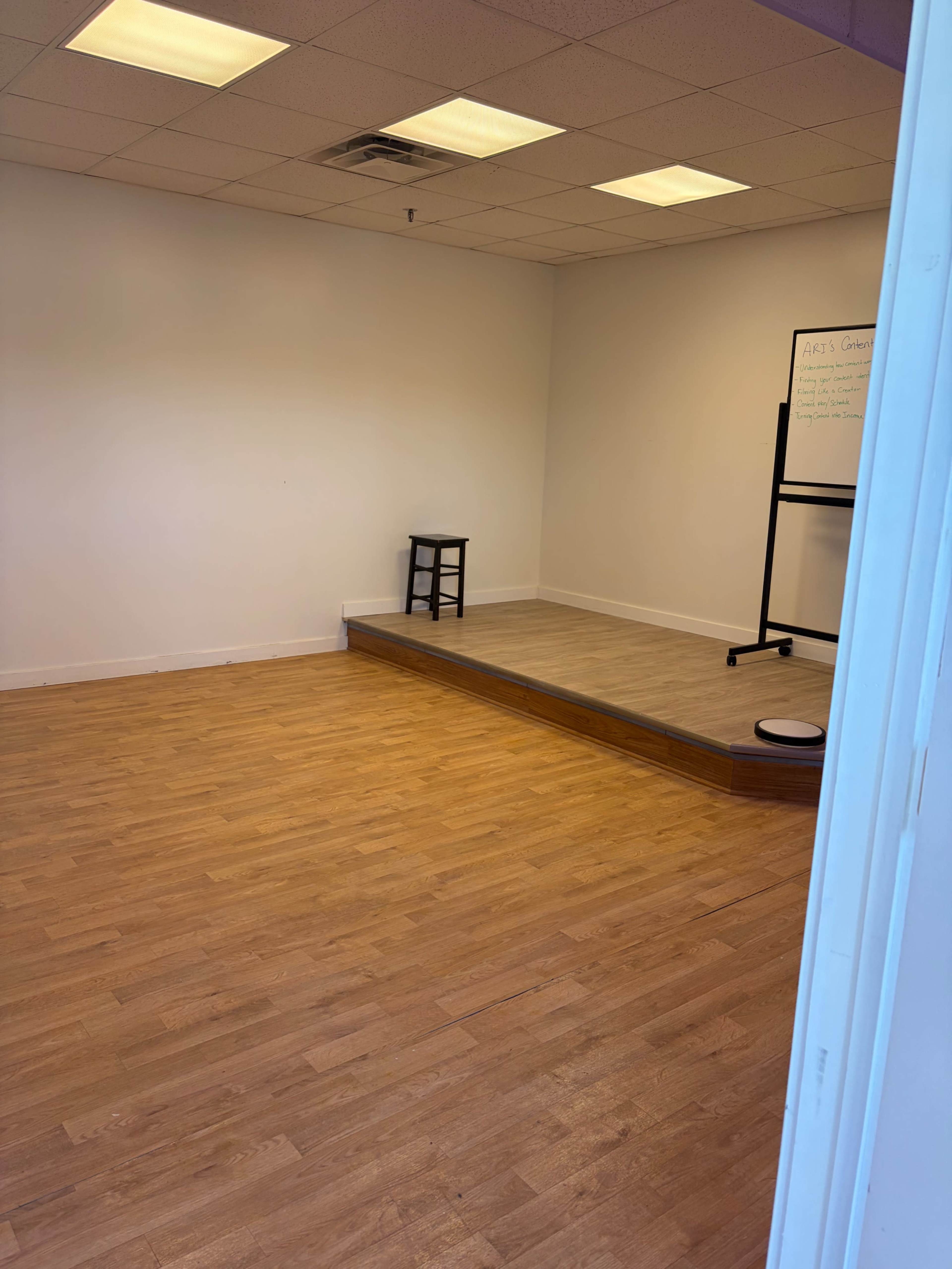 The image shows a spacious, empty room with wooden flooring, featuring a raised platform and a stool on one side.