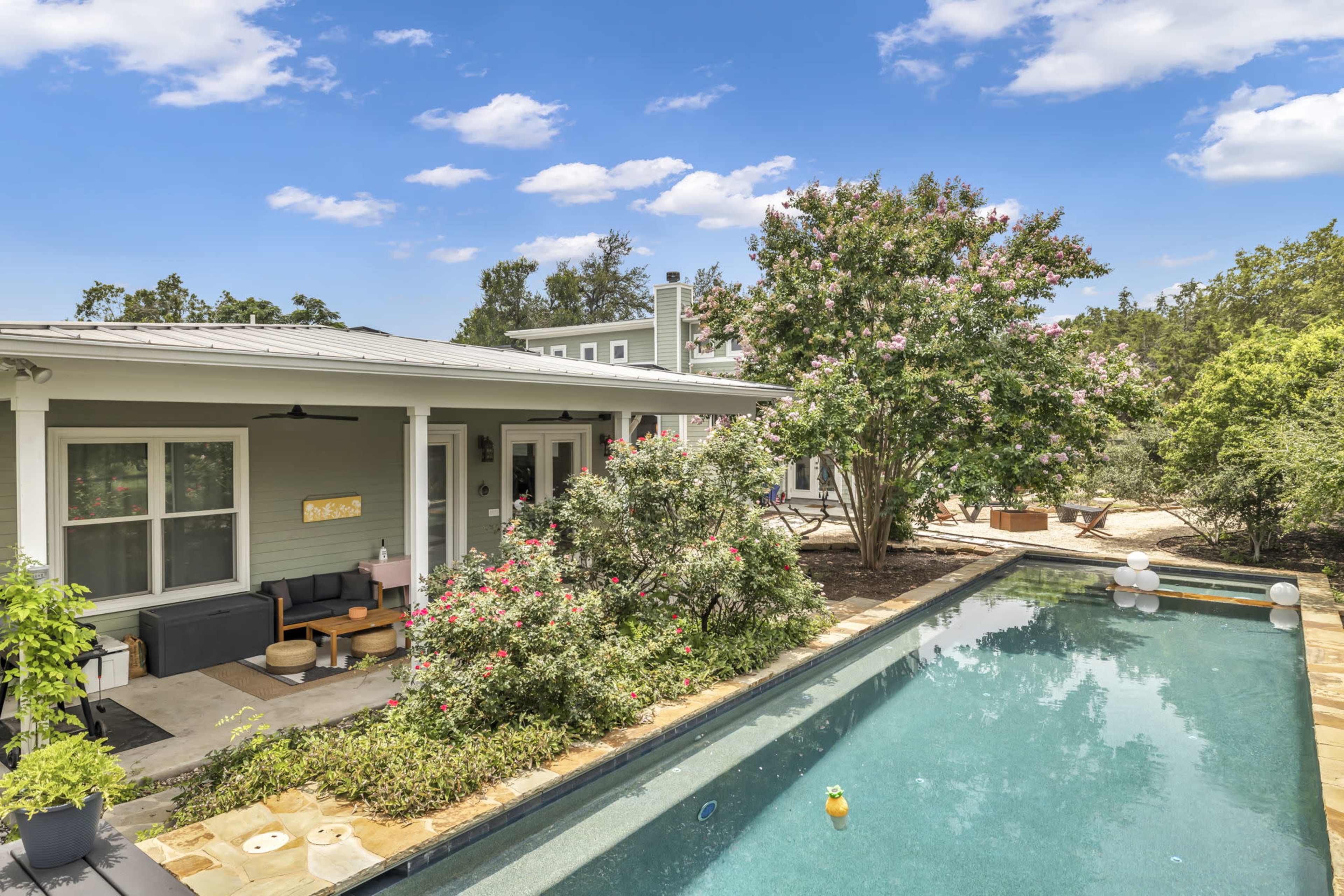A modern house with a swimming pool and landscaped garden features a patio area with seating and flowering trees in the background.