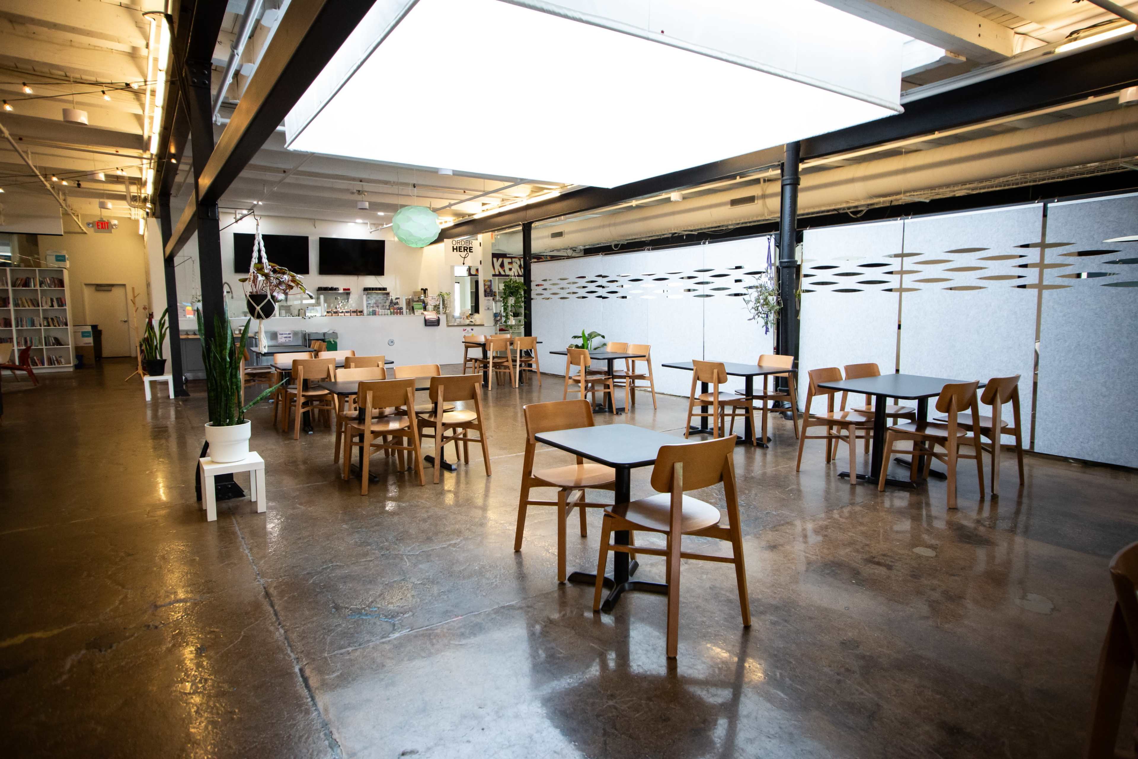 The image shows a spacious, modern café with wooden tables and chairs arranged across a polished concrete floor, featuring a counter and minimal decor in the background.