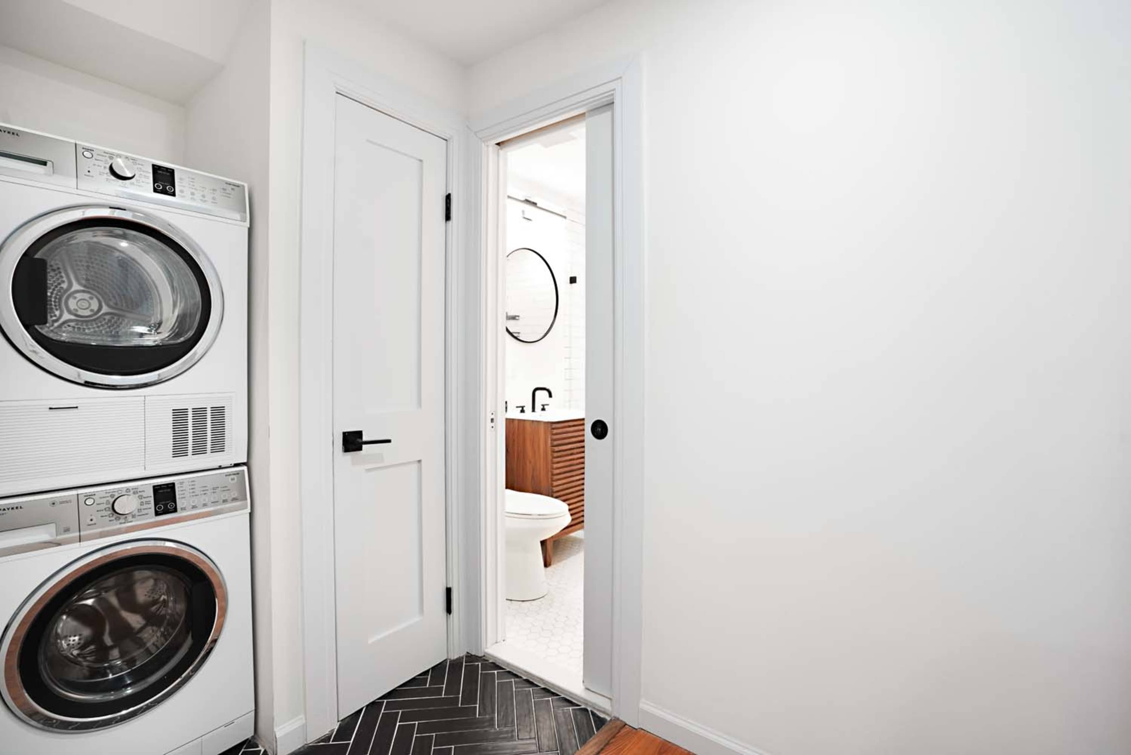 The image shows a laundry area with stacked washers and dryers next to a partially open door leading to a bathroom.