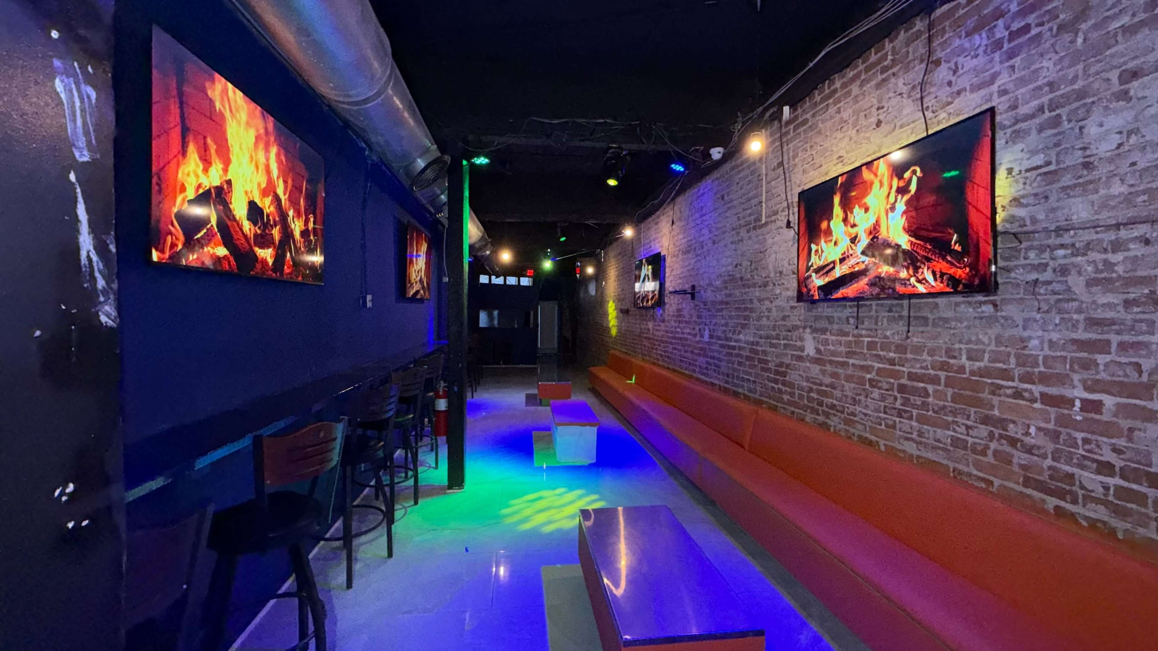 The image shows a dimly lit lounge with a long orange bench along a brick wall, illuminated by multicolored lights and featuring two large screens displaying fire imagery.