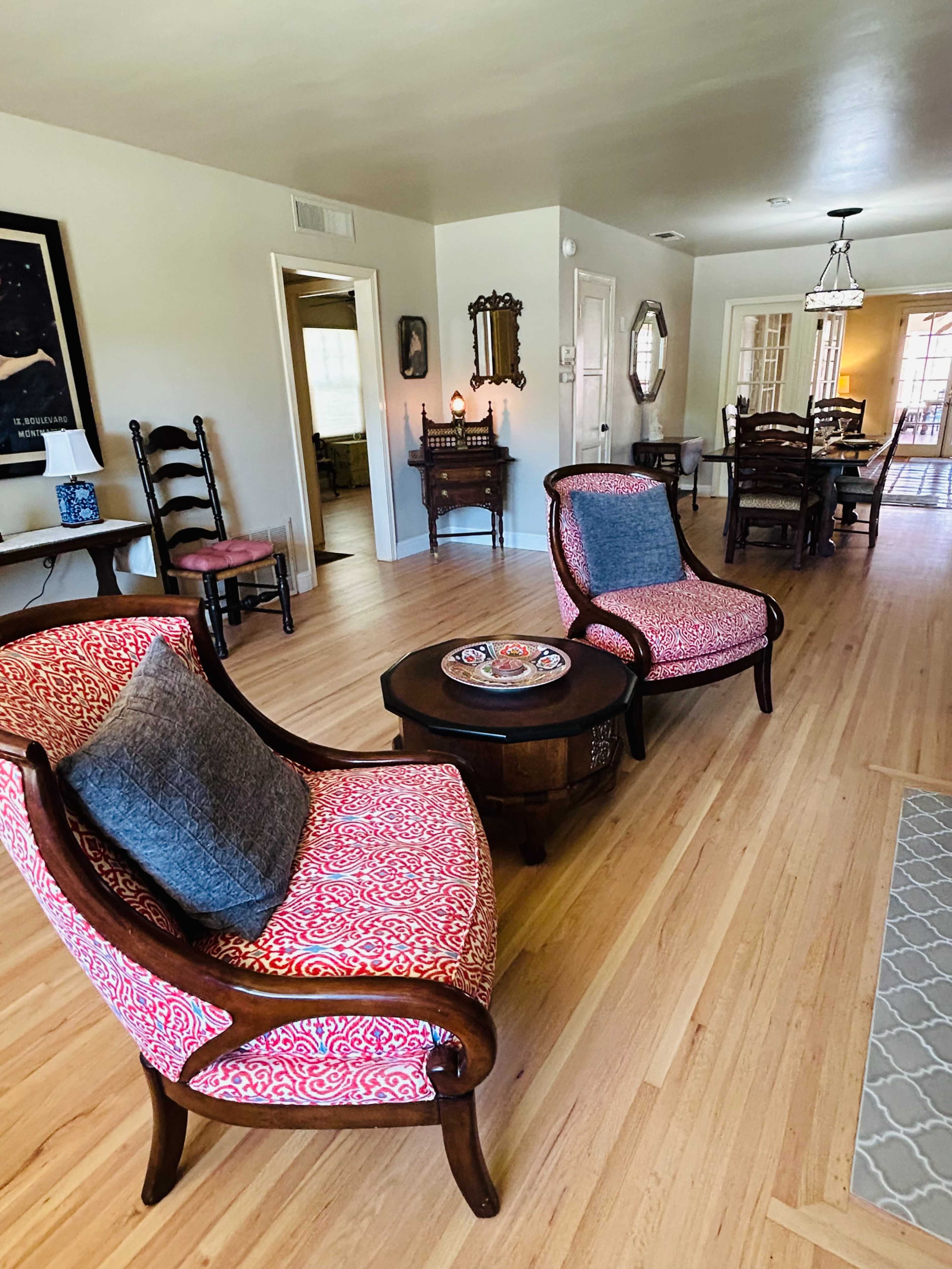 A living room furnished with patterned armchairs, a round wooden coffee table, and a dining area visible in the background.