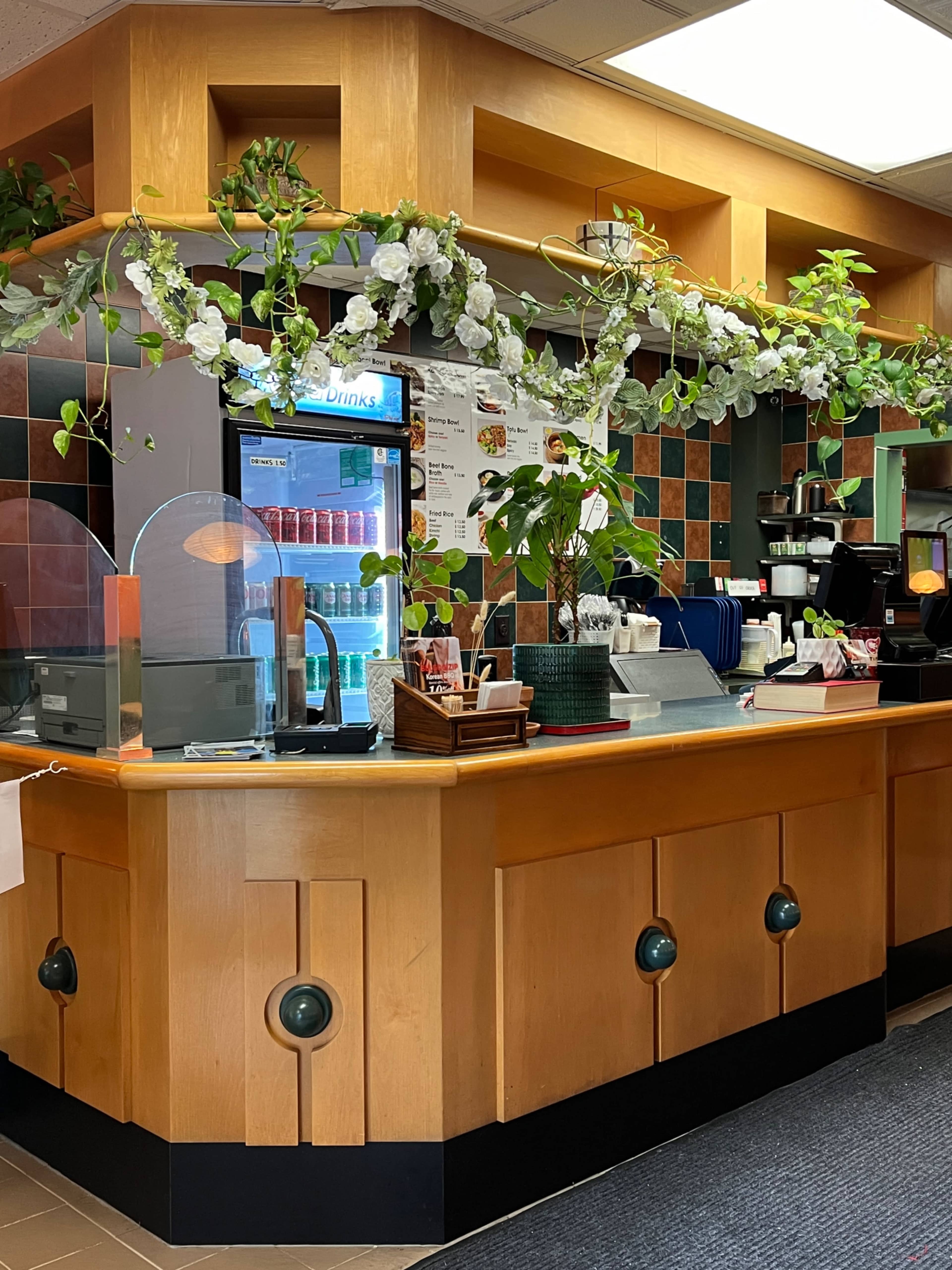A restaurant counter adorned with artificial flowers and a glass display refrigerator filled with drinks.