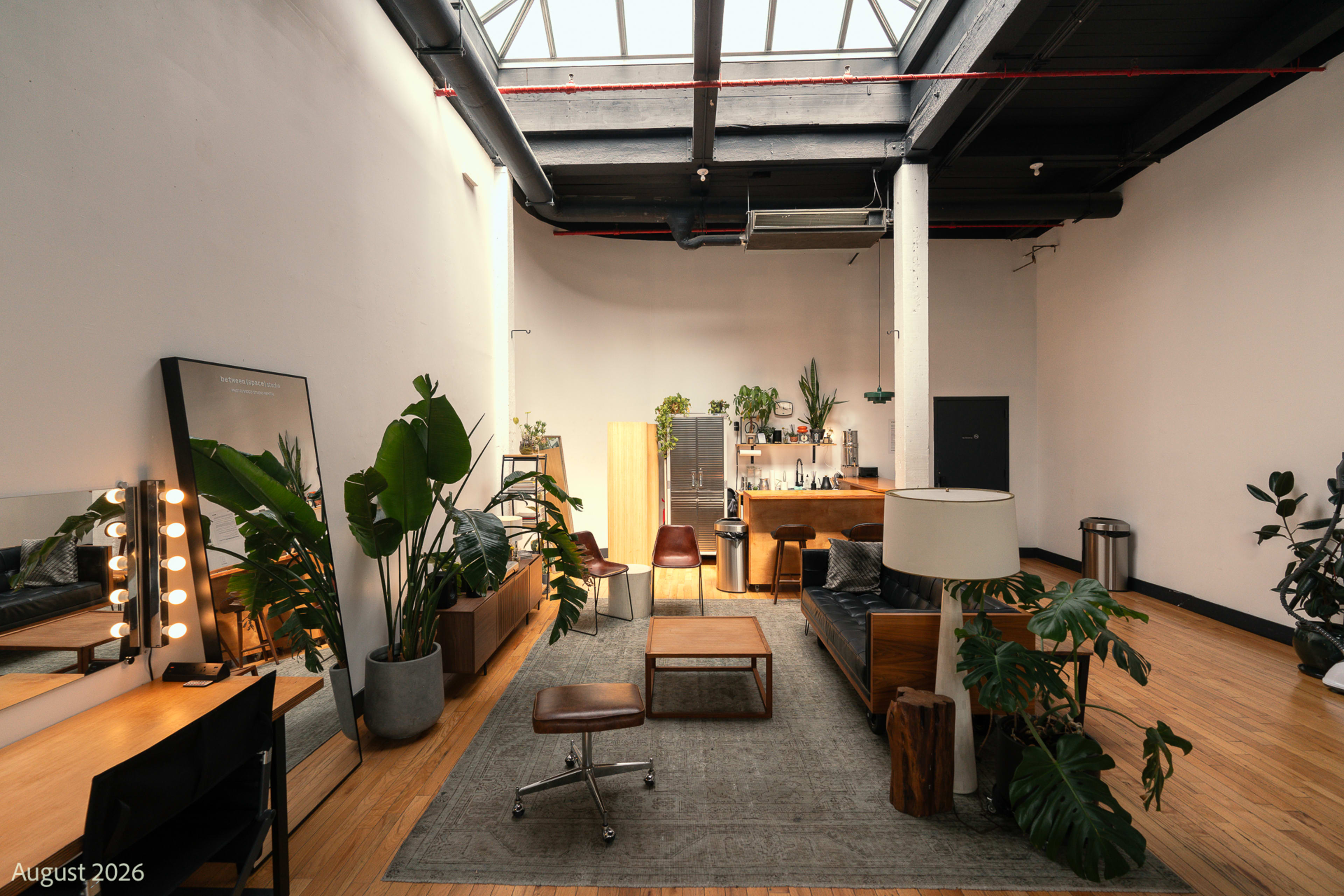 The image shows a spacious, well-lit room featuring a seating area with plants, a coffee table, and a bar setup against a minimalist backdrop.