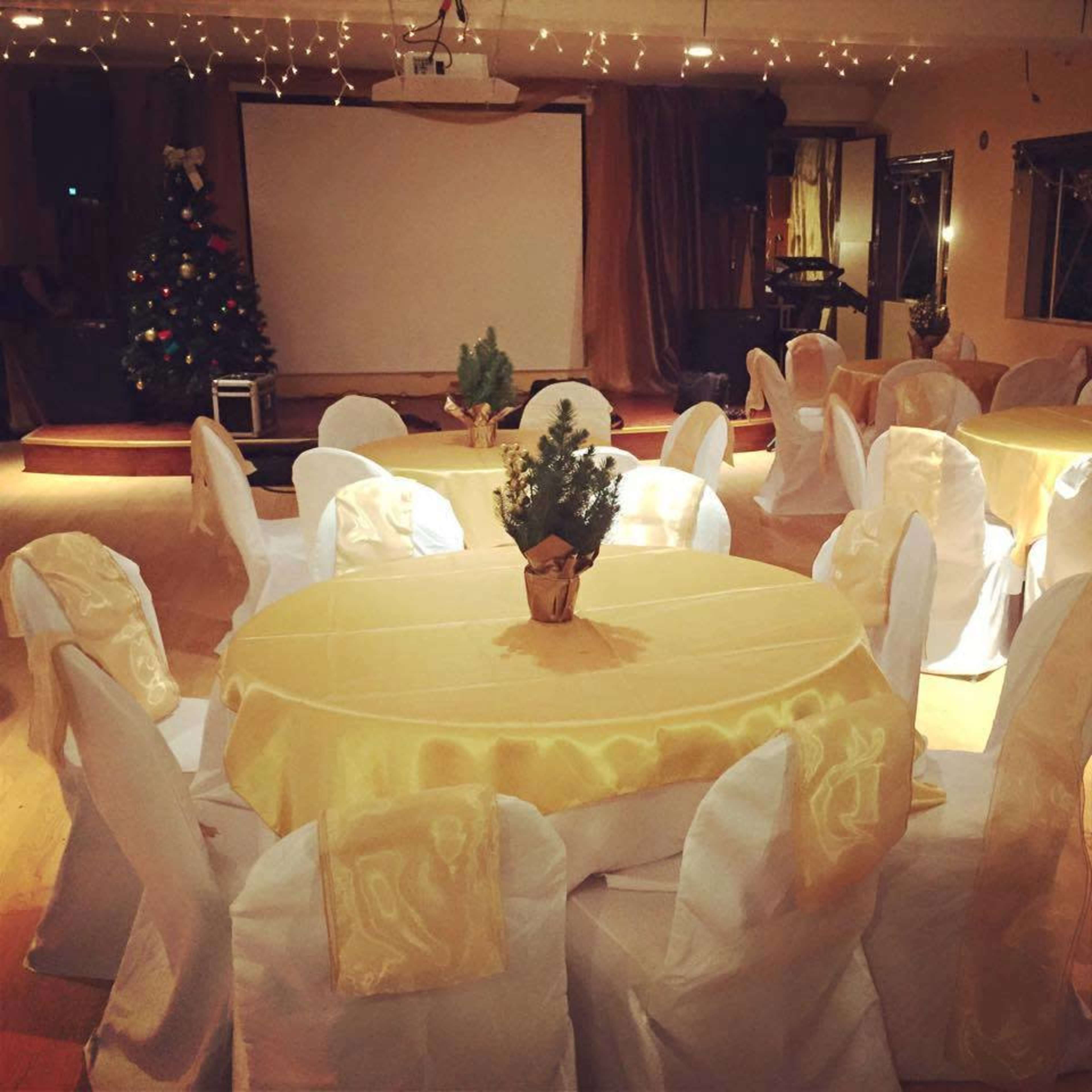 The image shows a banquet hall decorated with white tables and chairs, draped with yellow tablecloths, and a Christmas tree in the corner.