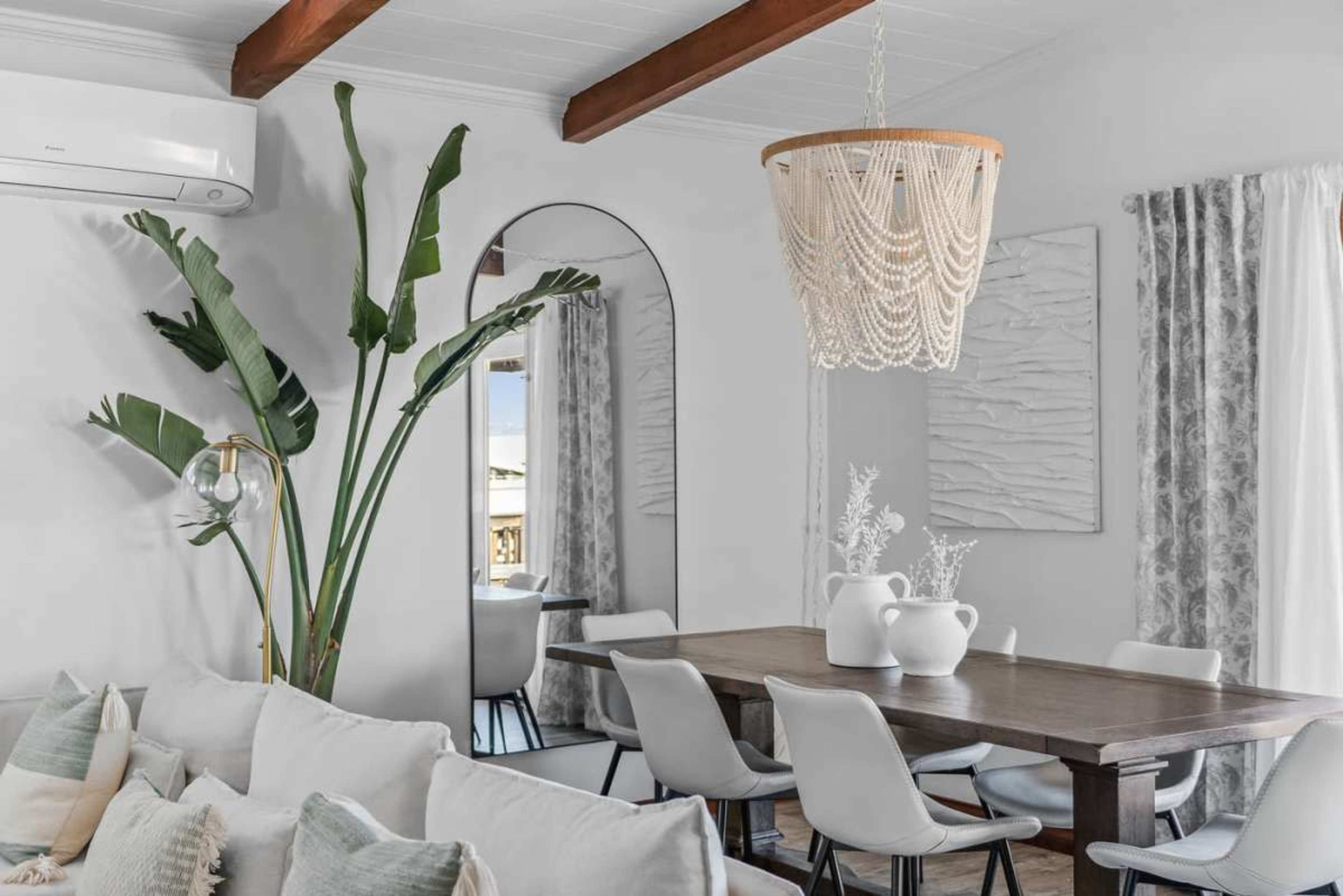 A modern dining area features a wooden table surrounded by white chairs, a large mirror, and a unique woven chandelier, with plants and decorative elements adding to the decor.