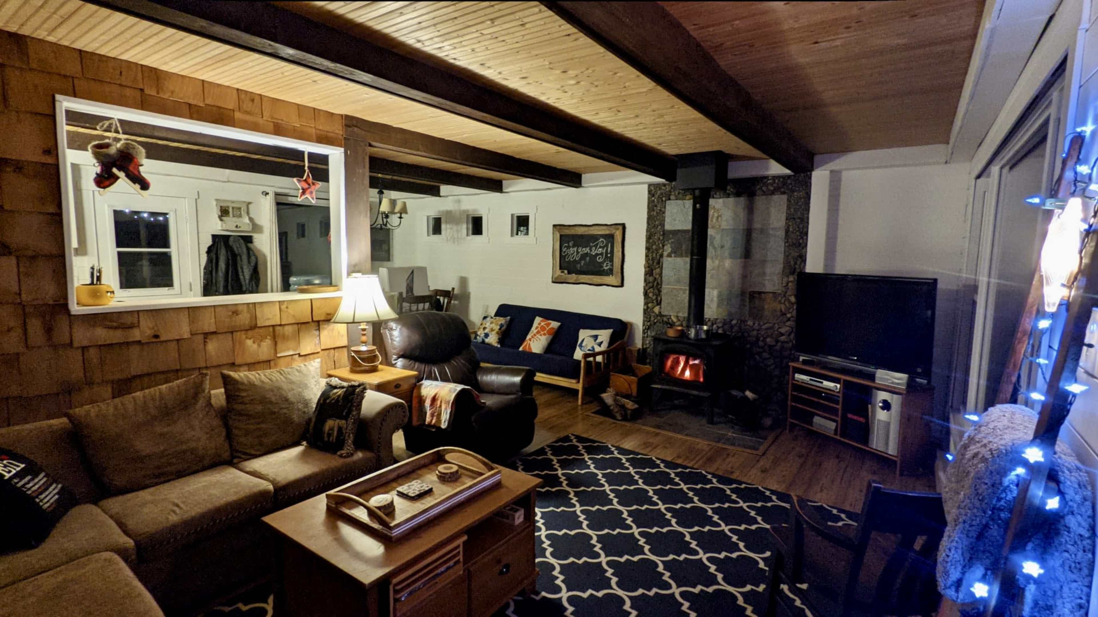 A cozy living room with a wooden-paneled wall, a comfortable seating area with a sofa and armchair, a coffee table, and a wood-burning stove surrounded by stone.