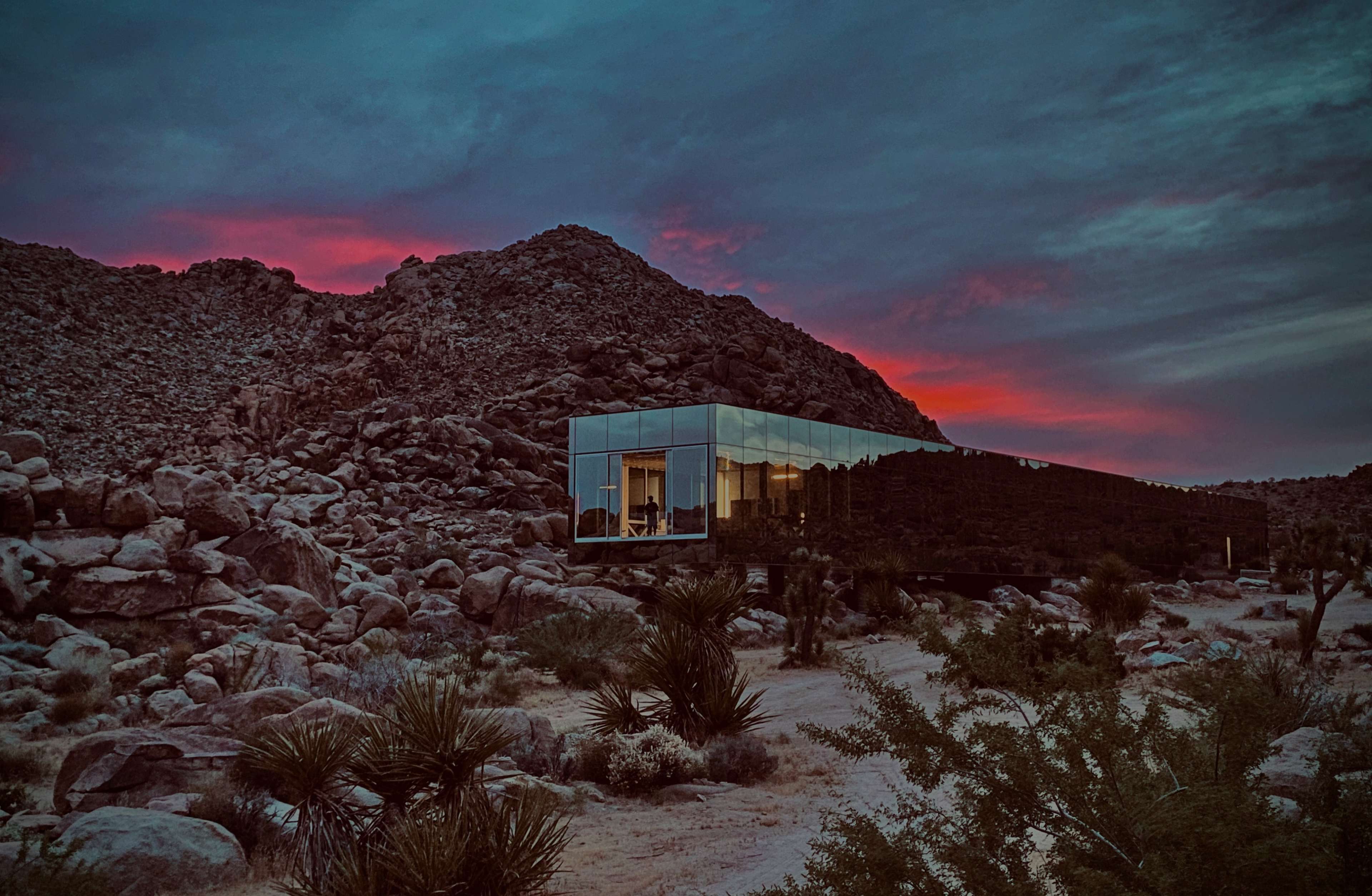 A modern glass house stands against a backdrop of rocky terrain and a colorful sunset in a desert landscape.