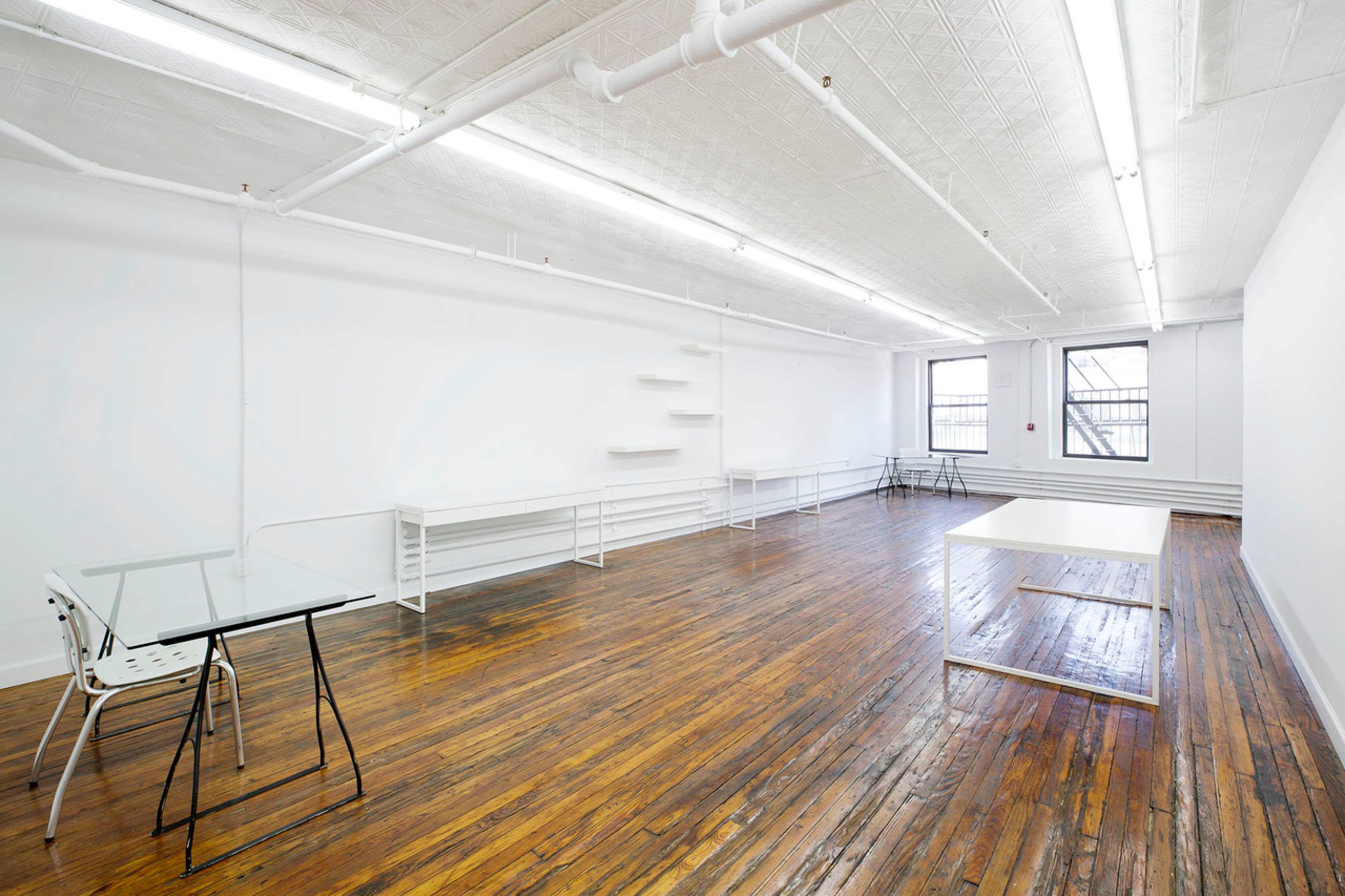 The image shows a spacious, empty room with hardwood floors, featuring white walls, overhead fluorescent lights, and minimal furniture including a glass table and a large white table.