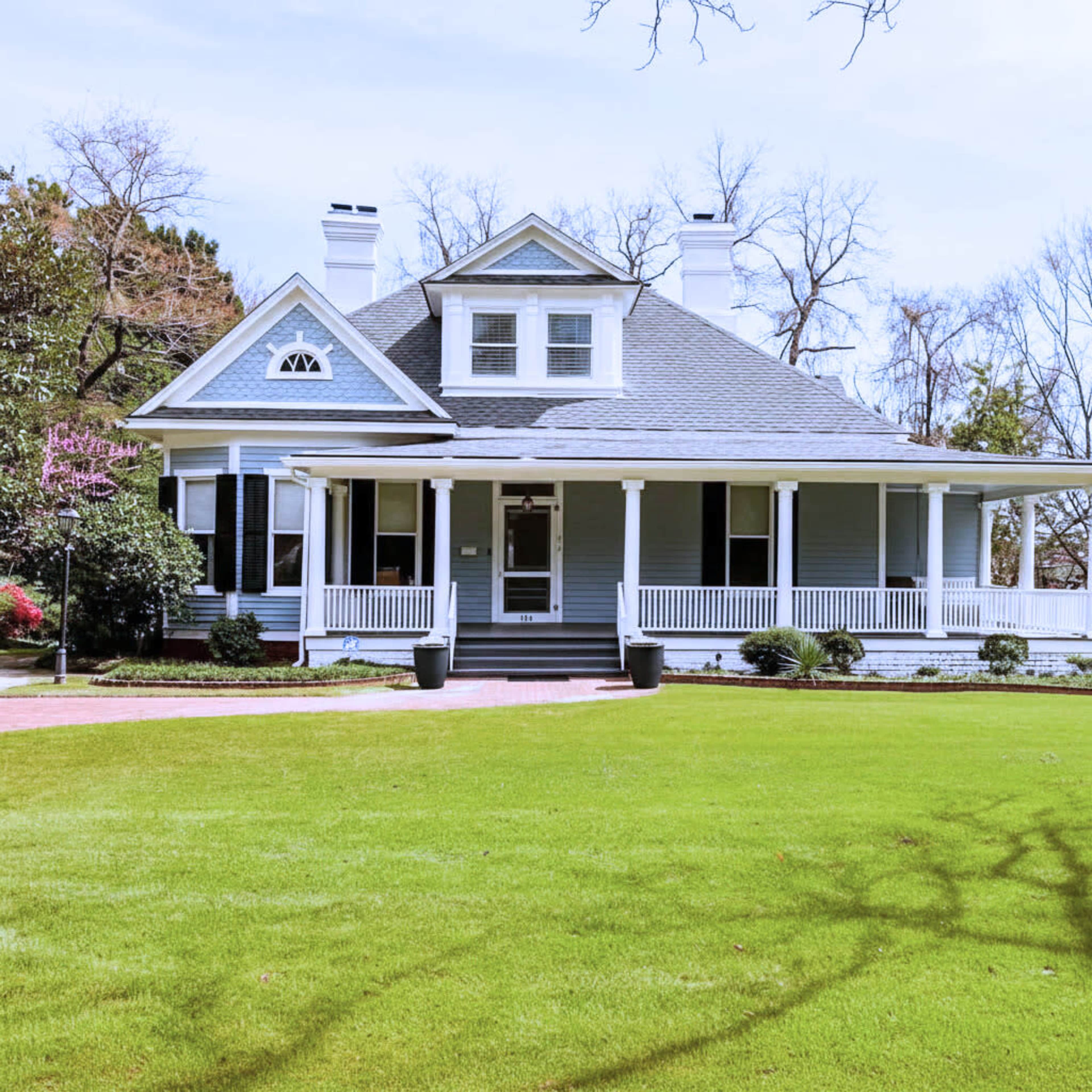 A light blue house with a peaked roof and a front porch is situated on a spacious green lawn.